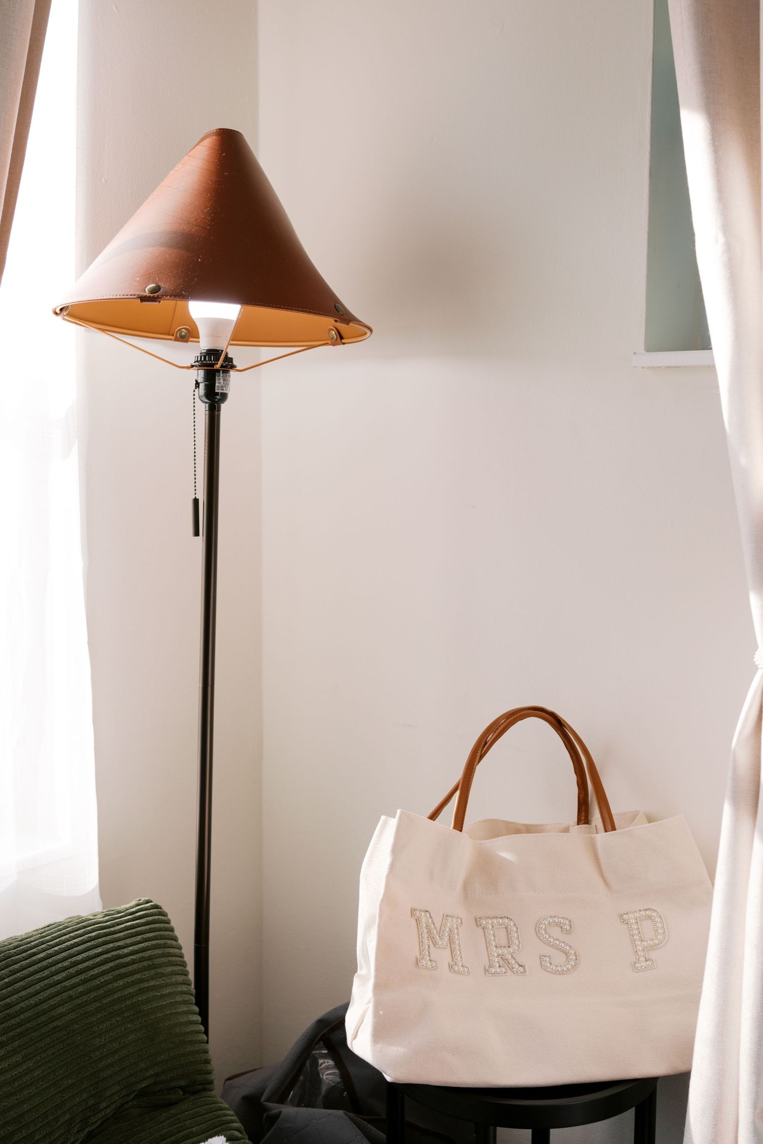 A white canvas tote bag labeled MRS P with leather handles sits beside a brown conical floor lamp near white curtains.