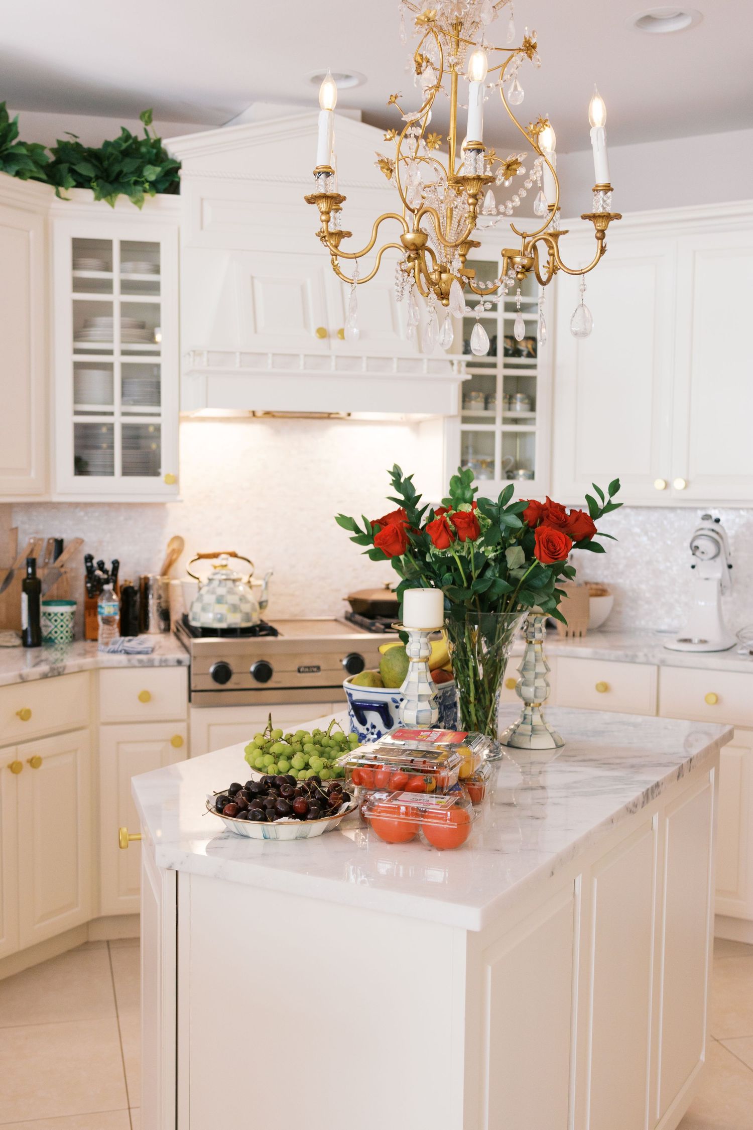 Elegant white kitchen with gold chandelier, marble island, red roses, fresh fruit, and white cabinetry with brass hardware.