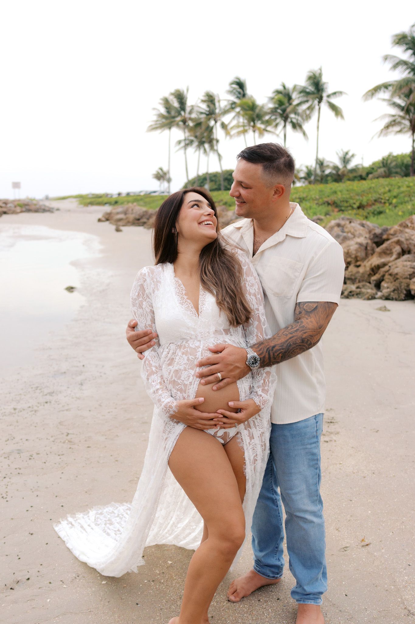 Pregnant couple embracing on a tropical beach, woman in white lace maternity gown, man in white shirt and jeans.