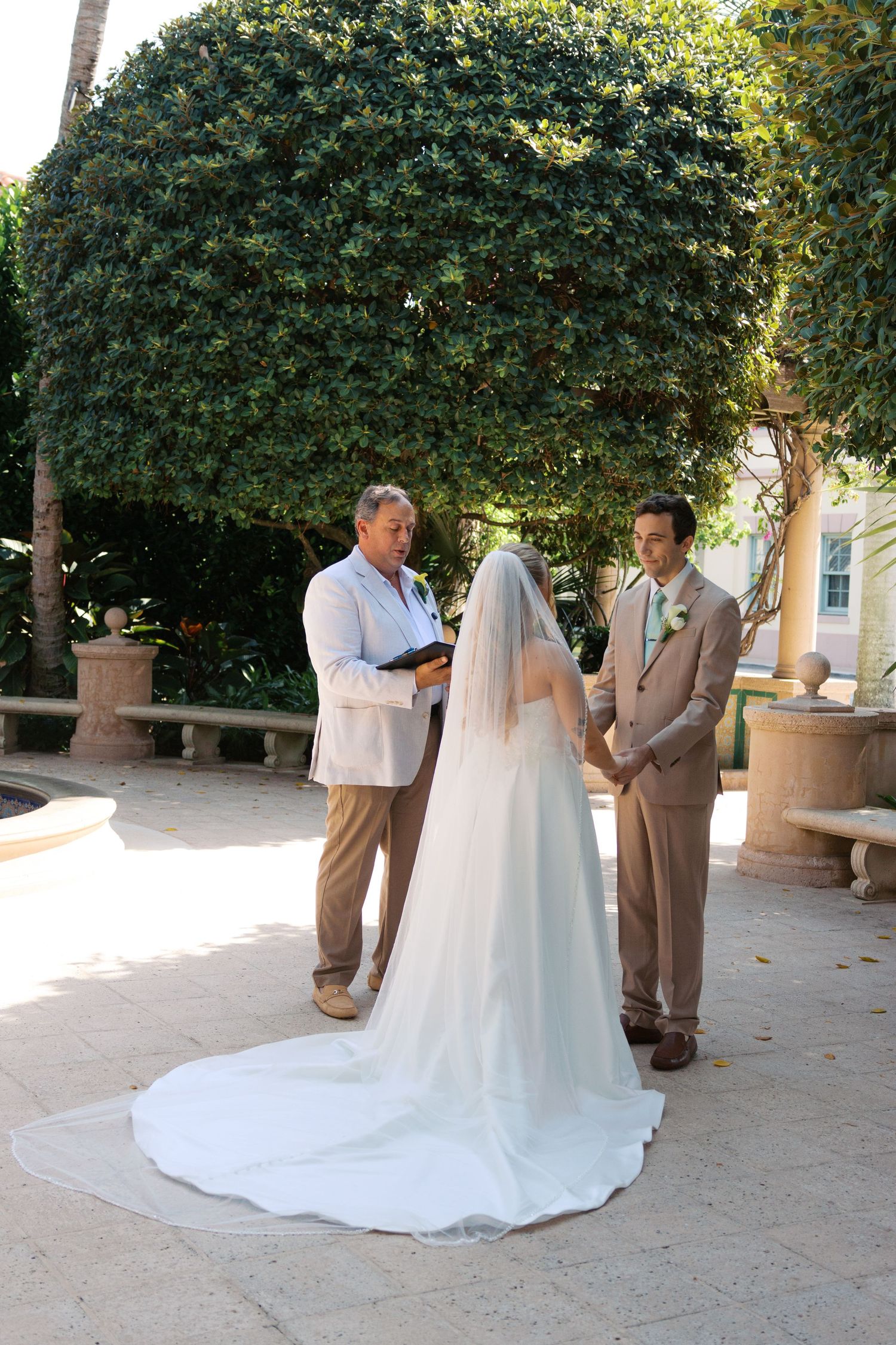 Outdoor wedding ceremony with bride and groom exchanging vows before officiant, lush green hedges backdrop, sunny day.