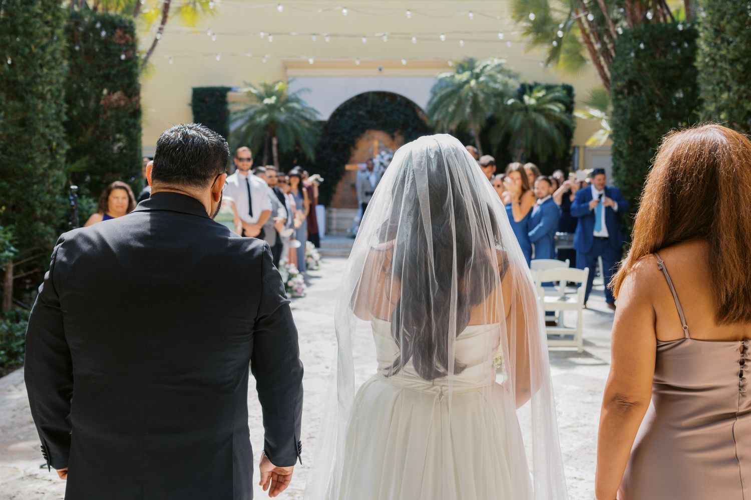 Bride in white gown and veil walks with groom in black suit toward outdoor wedding ceremony guests.