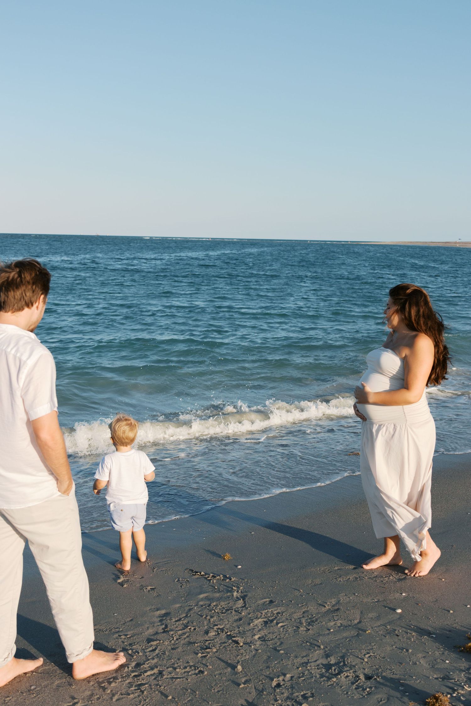 Family of three at beach shoreline, pregnant mother in white dress holds belly as toddler approaches ocean waves.