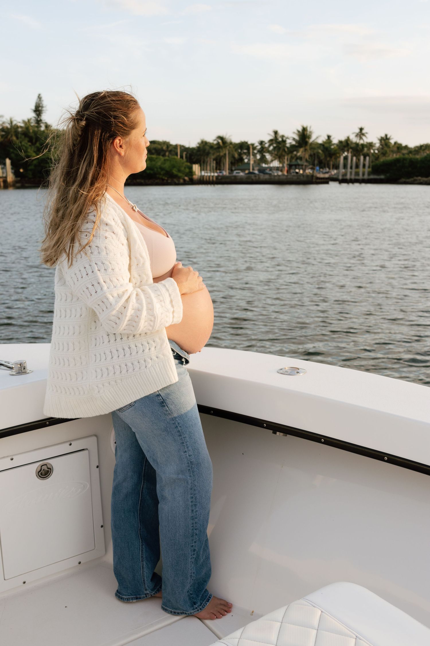 Side view of an expectant mother in a cream cardigan sitting on a boat with waterfront scenery.