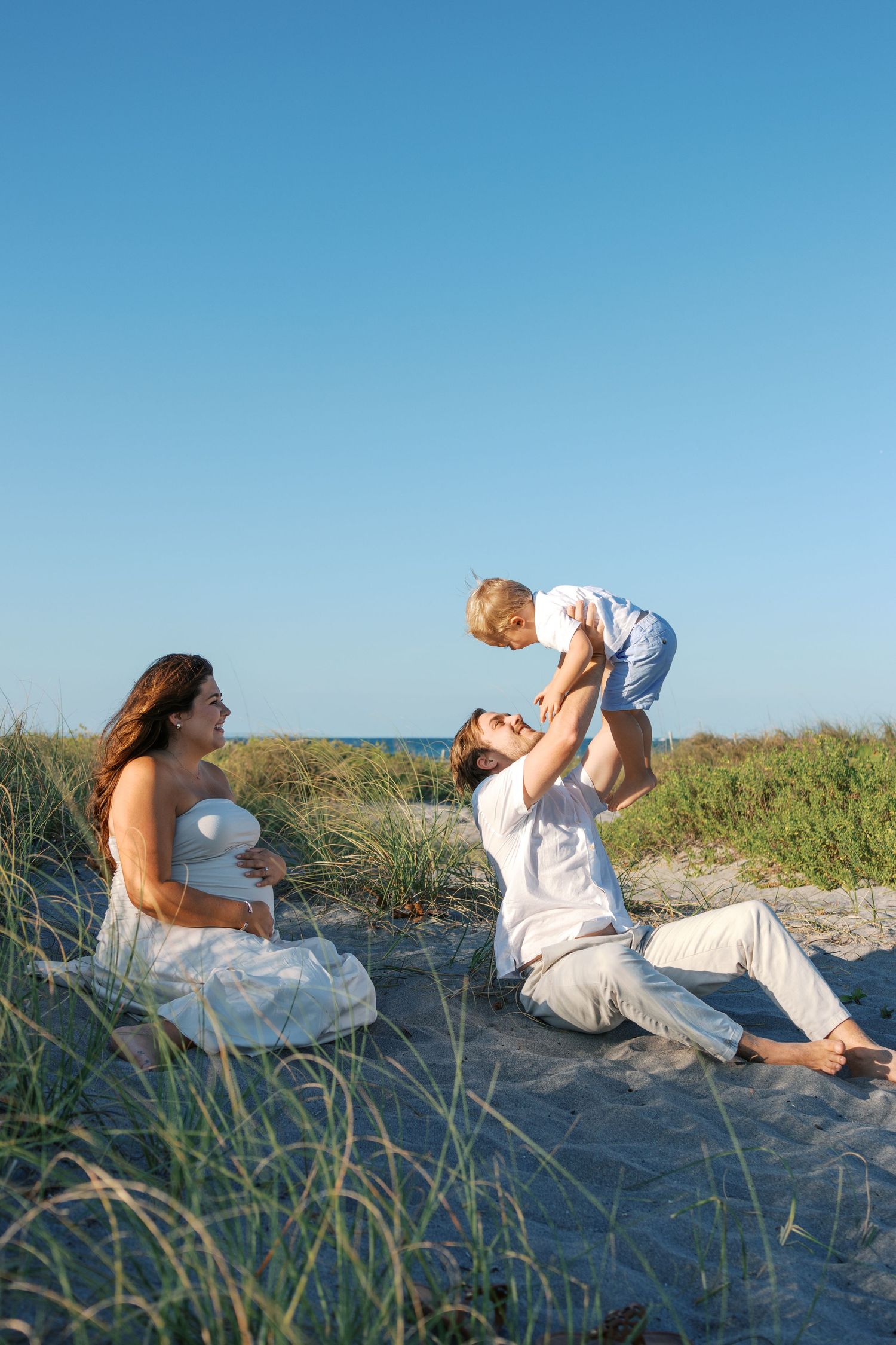 Happy father lifting toddler son in air on beach dunes while pregnant mother watches and smiles nearby.