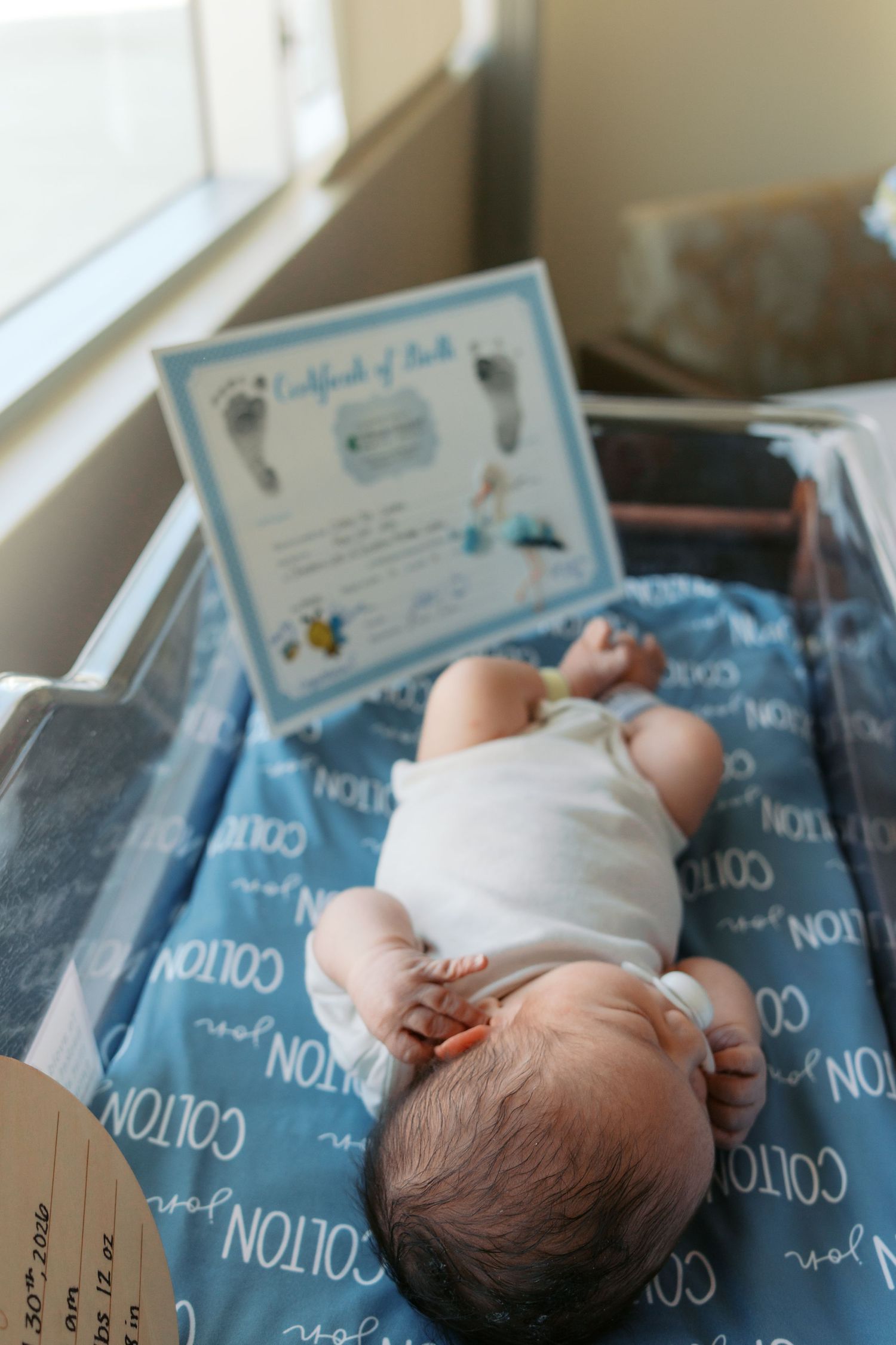 Newborn baby lying on personalized blue blanket named Colton next to a Certificate of Birth in hospital.