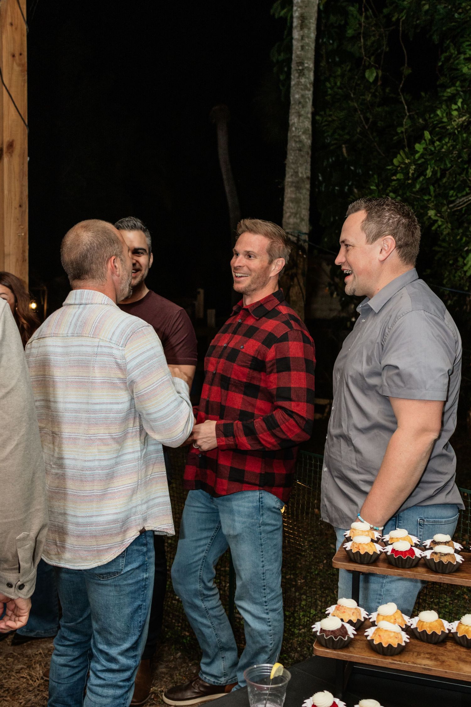 Men socializing at an outdoor evening gathering near a dessert display with cupcakes and pastries.