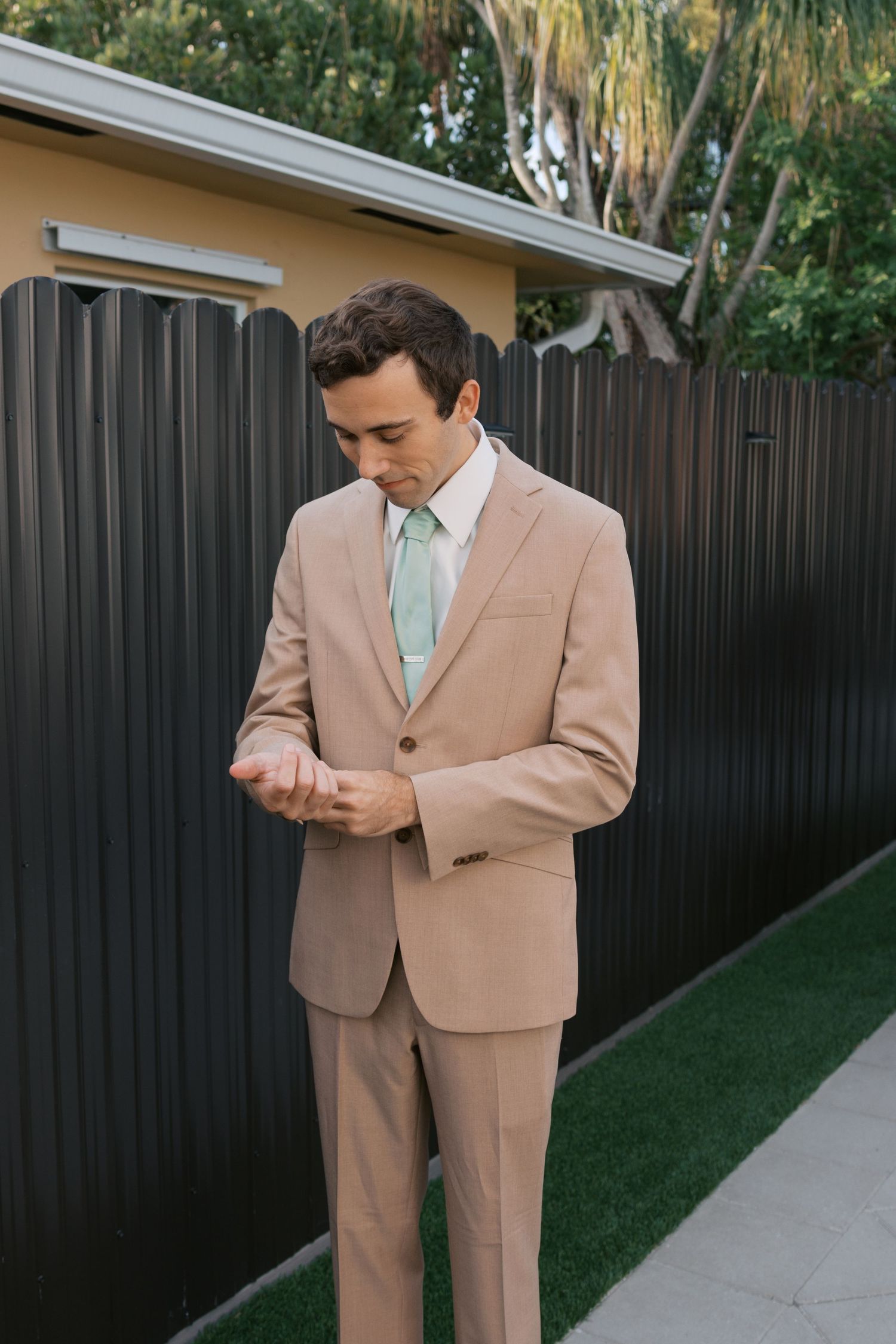 A man in a tan suit and mint green tie adjusts his cufflinks outdoors near a dark fence.