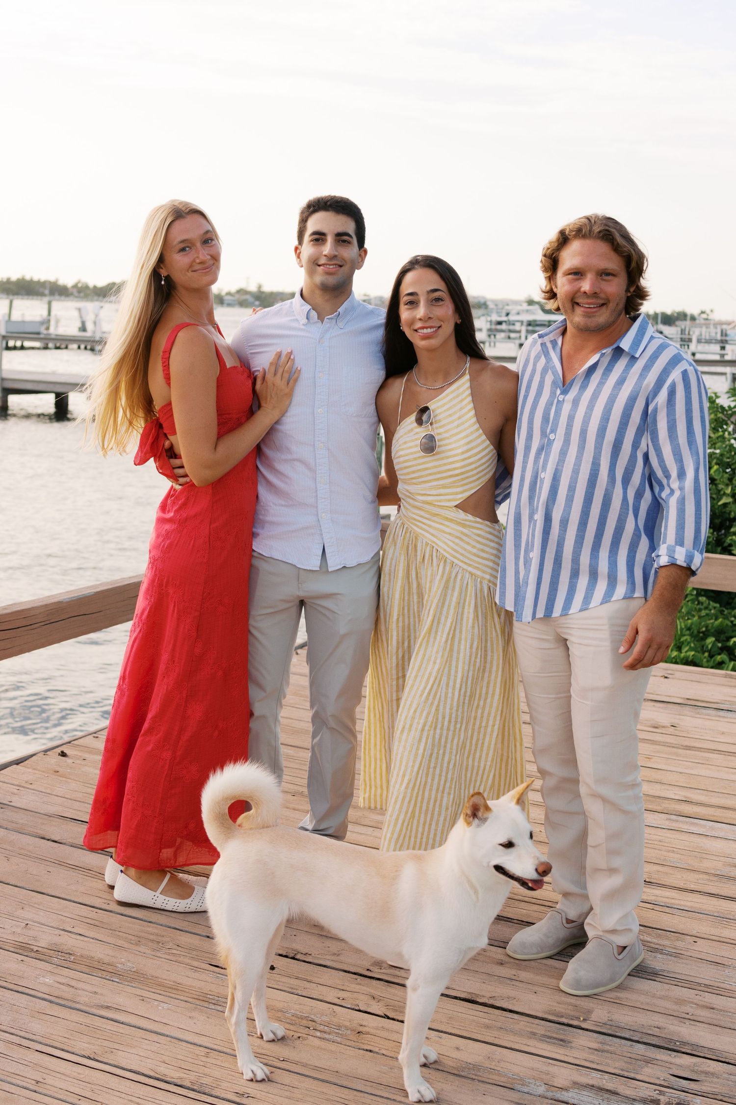 Four people and a white Shiba Inu dog pose on a waterfront dock at sunset, dressed in colorful summer attire.