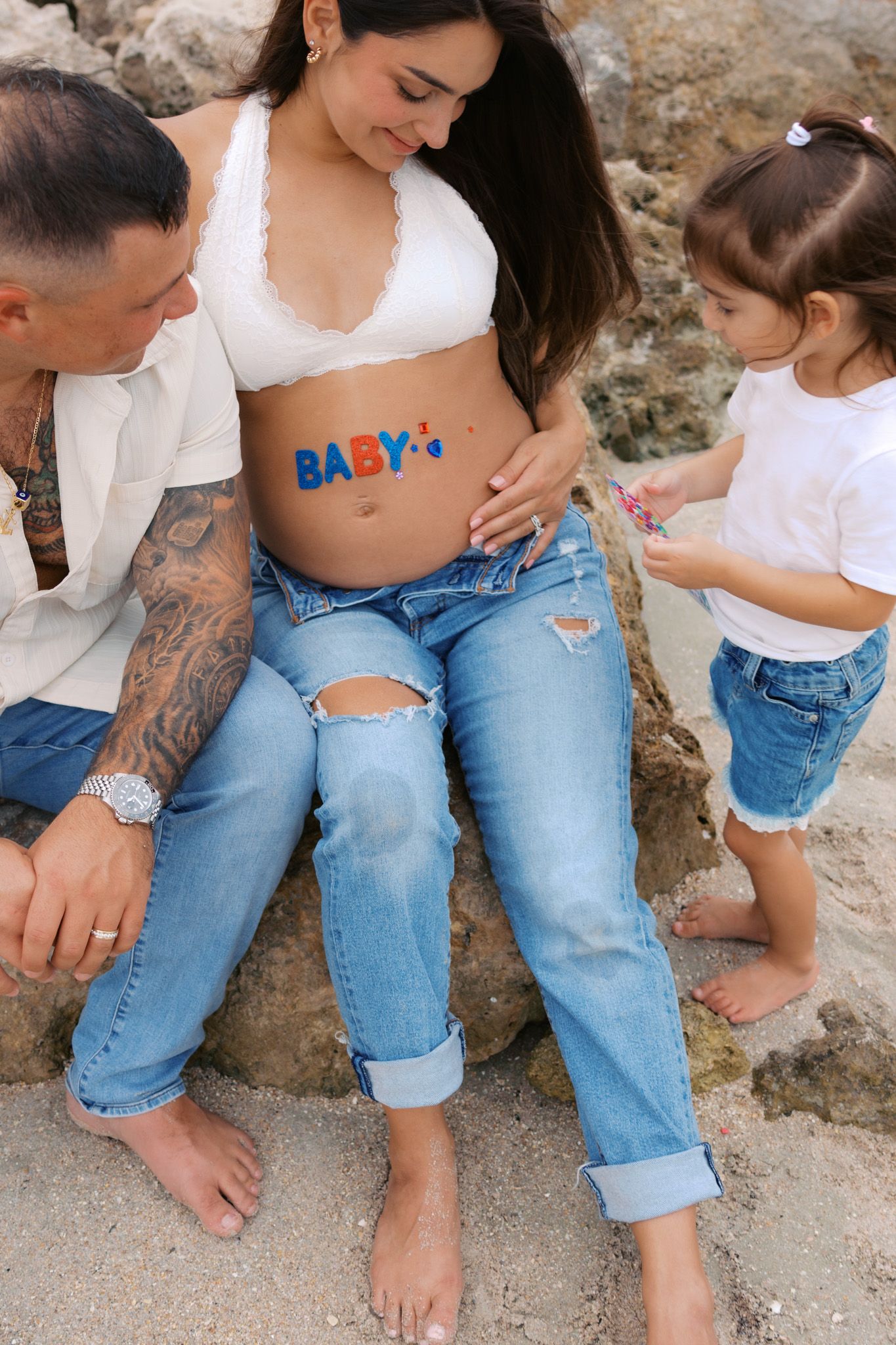 Family maternity photo with pregnant mom, tattooed dad, and young daughter touching painted baby bump outdoors.