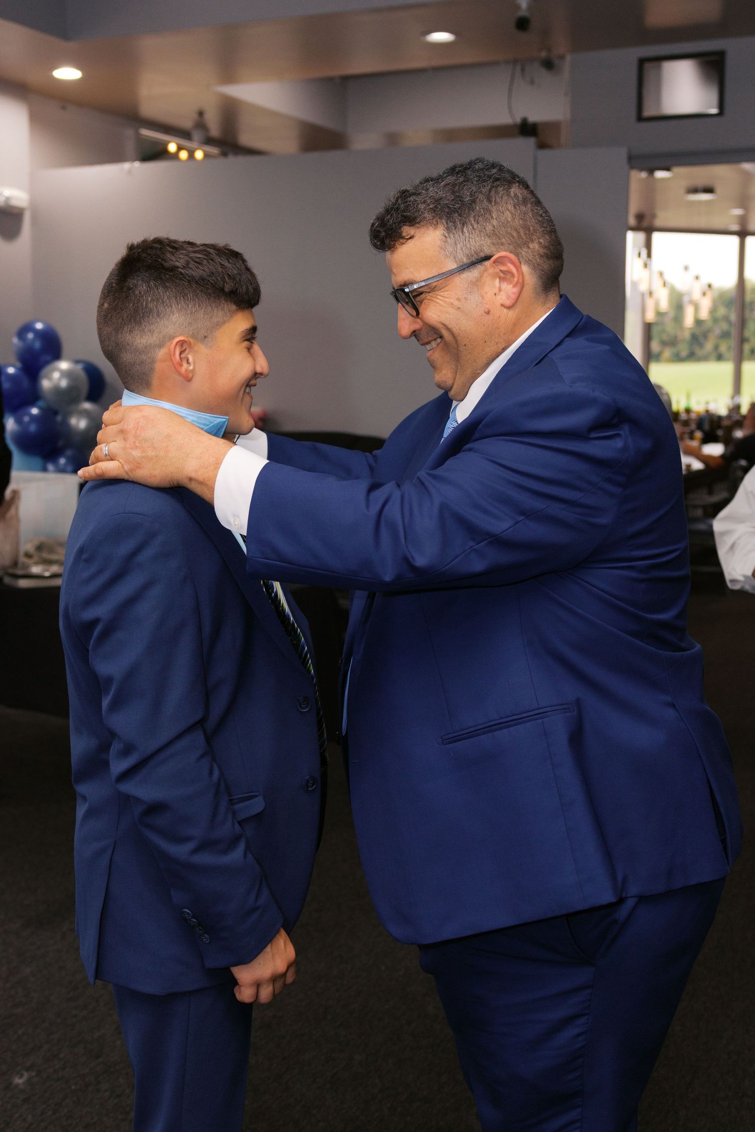 Man in blue suit adjusting boy's tie at formal celebration event with blue balloon decorations.