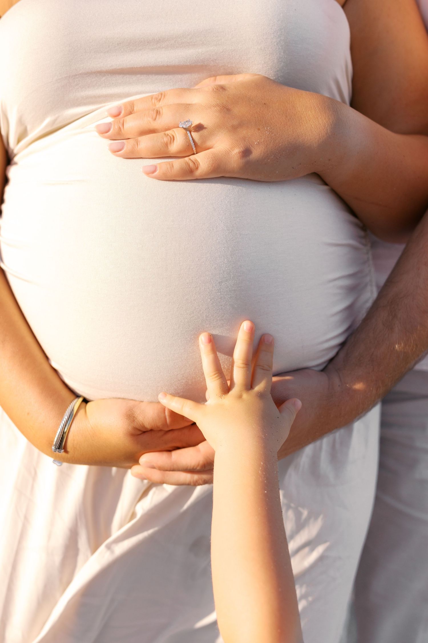 A child's hand touches a pregnant woman's belly while she gently cradles it, wearing a white dress in warm sunlight.