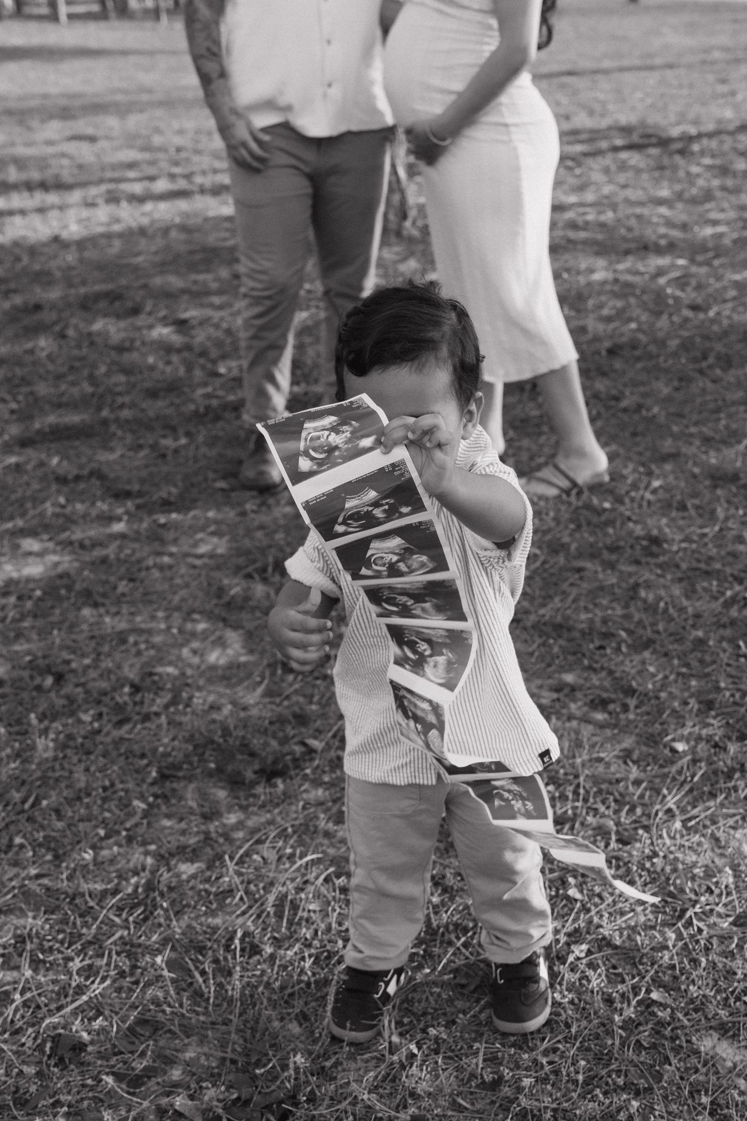 Toddler holding ultrasound photos outdoors in black and white maternity family photo session.