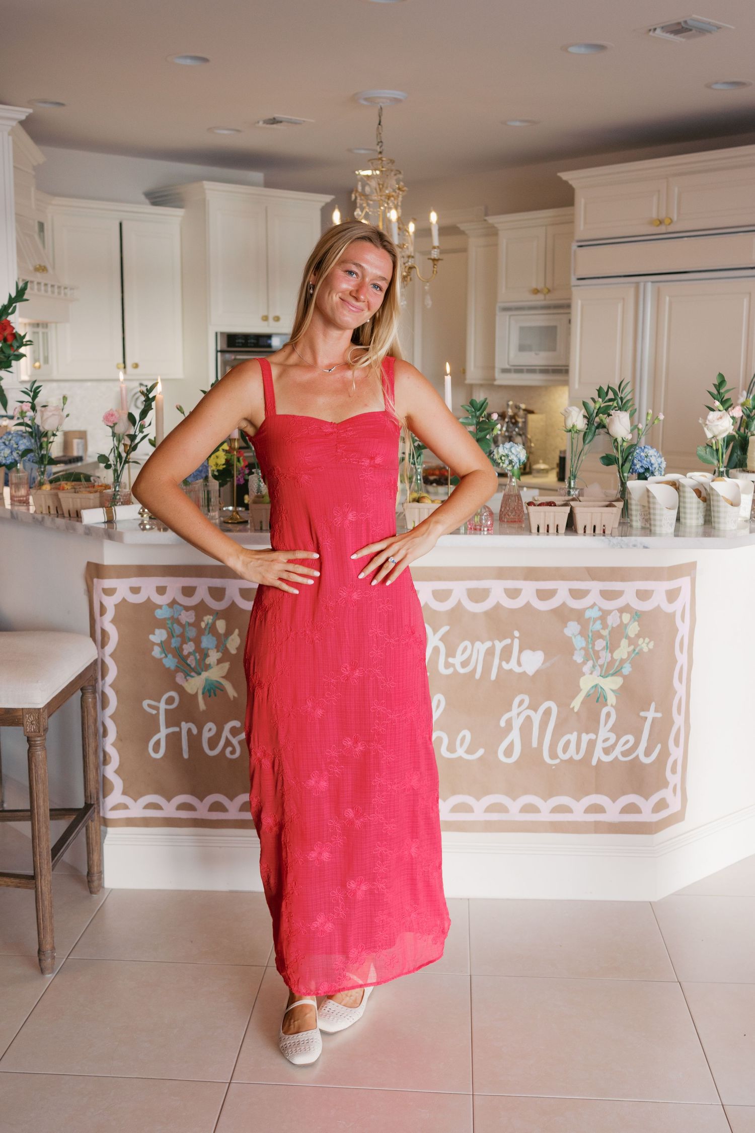 Woman in red maxi dress posing in elegant kitchen with floral decorations and market-themed banner.