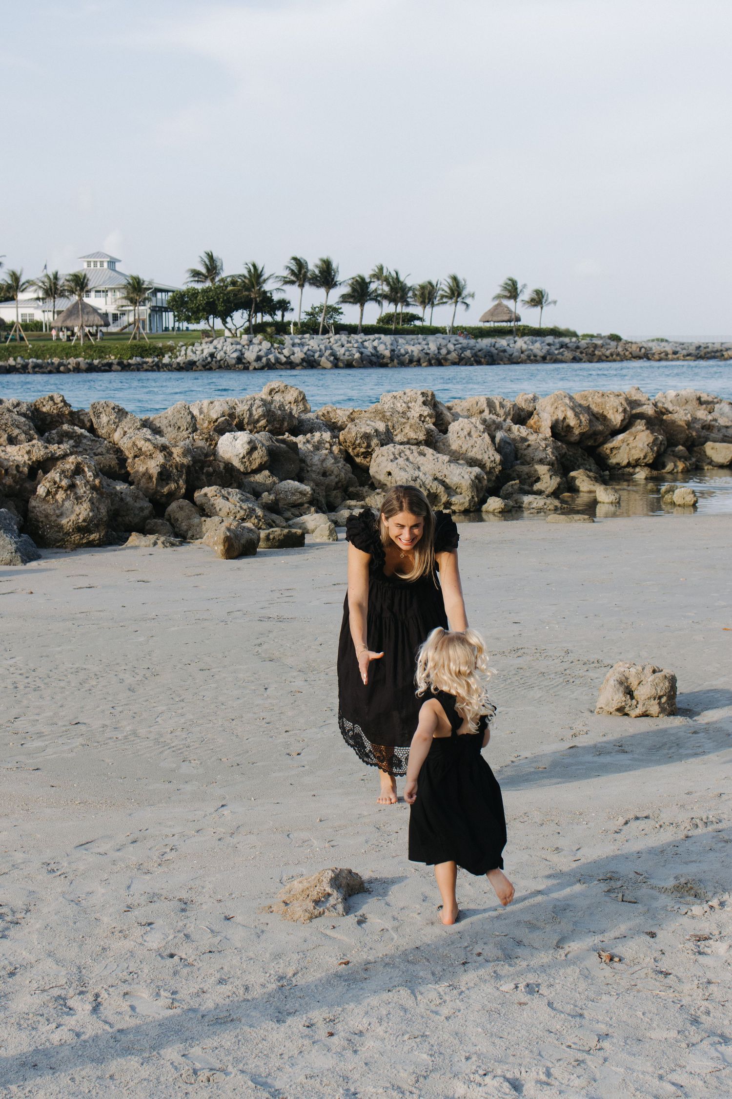 Two people in black dresses walking on beach with rocky shoreline and palm trees.