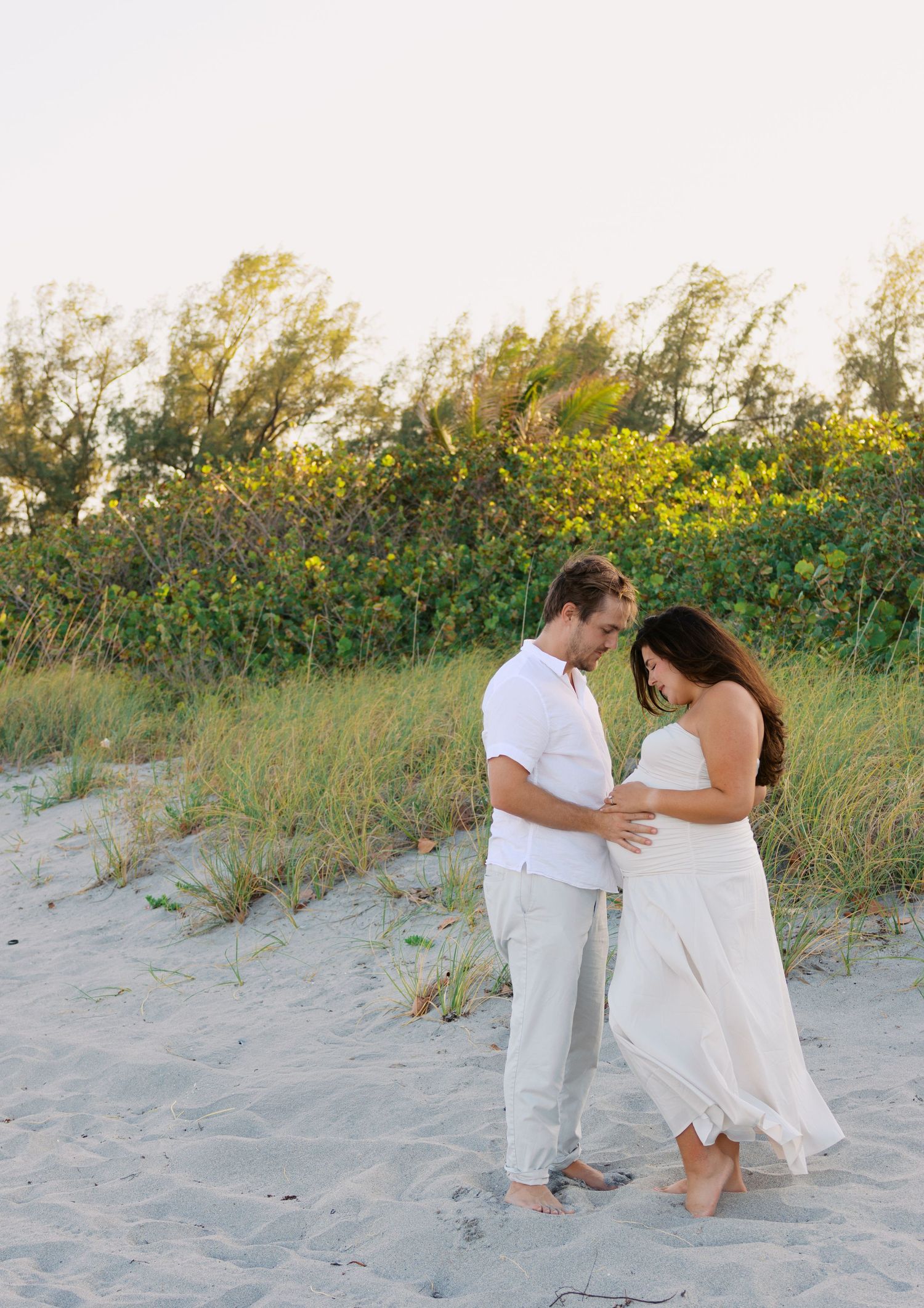 Couple in white outfits share tender moment on sandy beach, woman visibly pregnant, surrounded by dune grass.