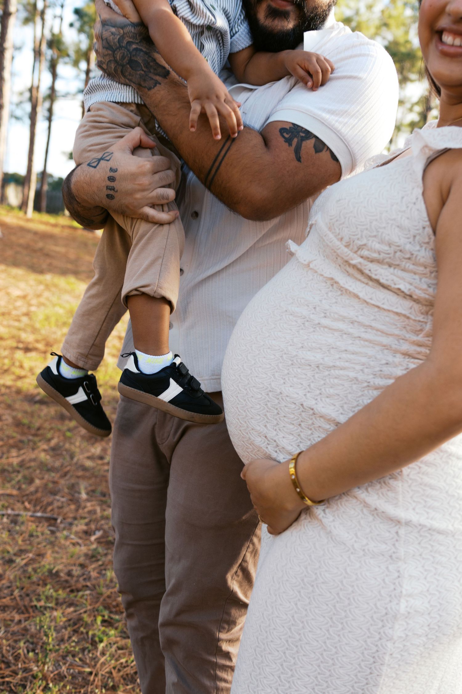 Pregnant woman in white dress stands with tattooed partner holding toddler outdoors among pine trees.