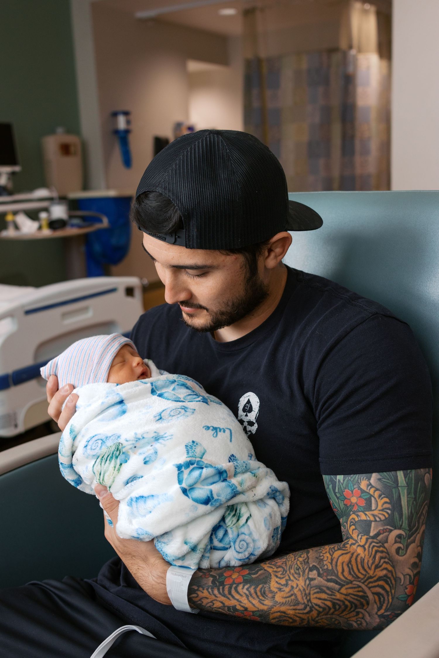 Tattooed father in black cap tenderly holding his swaddled newborn baby in a hospital room.