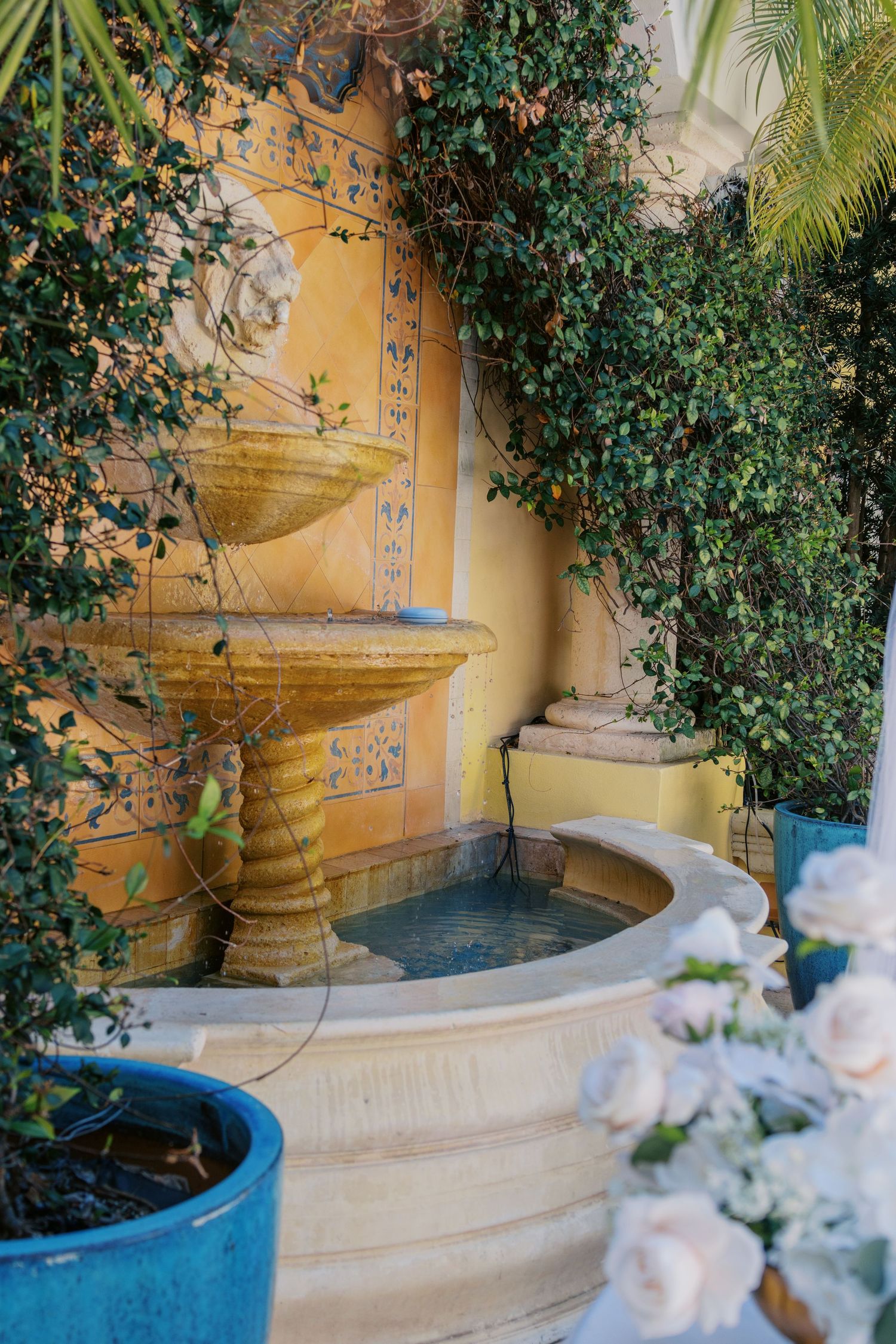 A tiered stone fountain surrounded by lush green ivy against a yellow tiled wall in a serene garden courtyard.