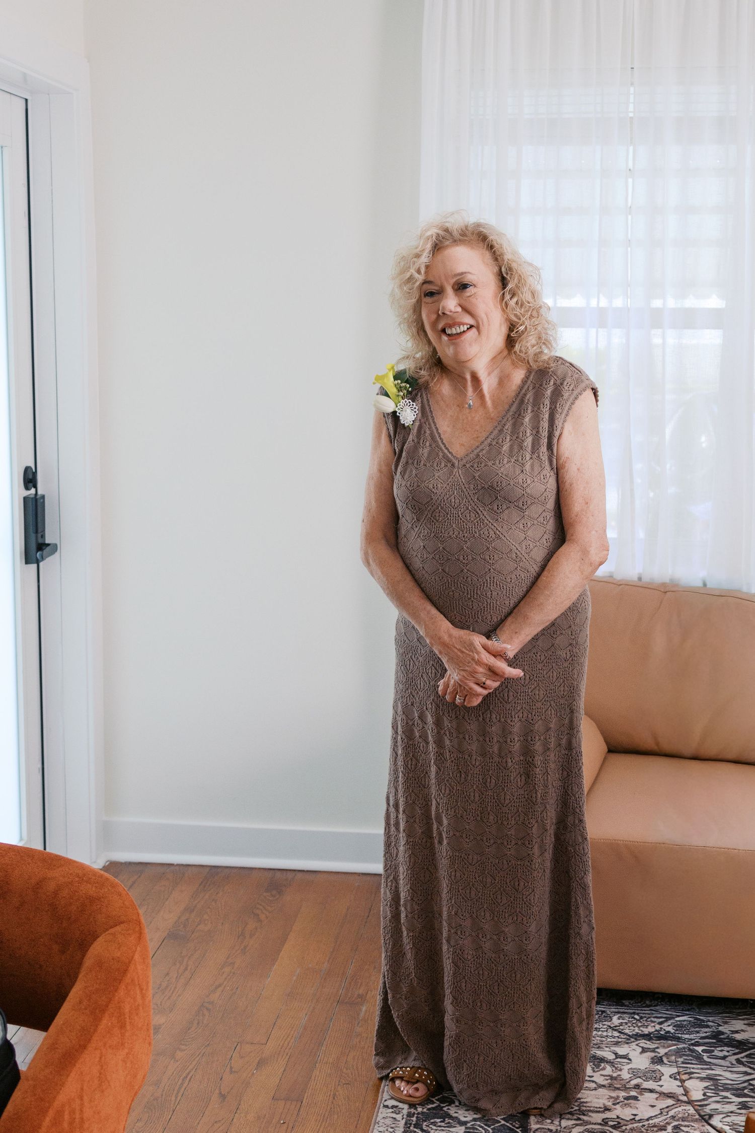 Elegant older woman in a brown maxi dress with floral corsage smiling indoors at a formal event.