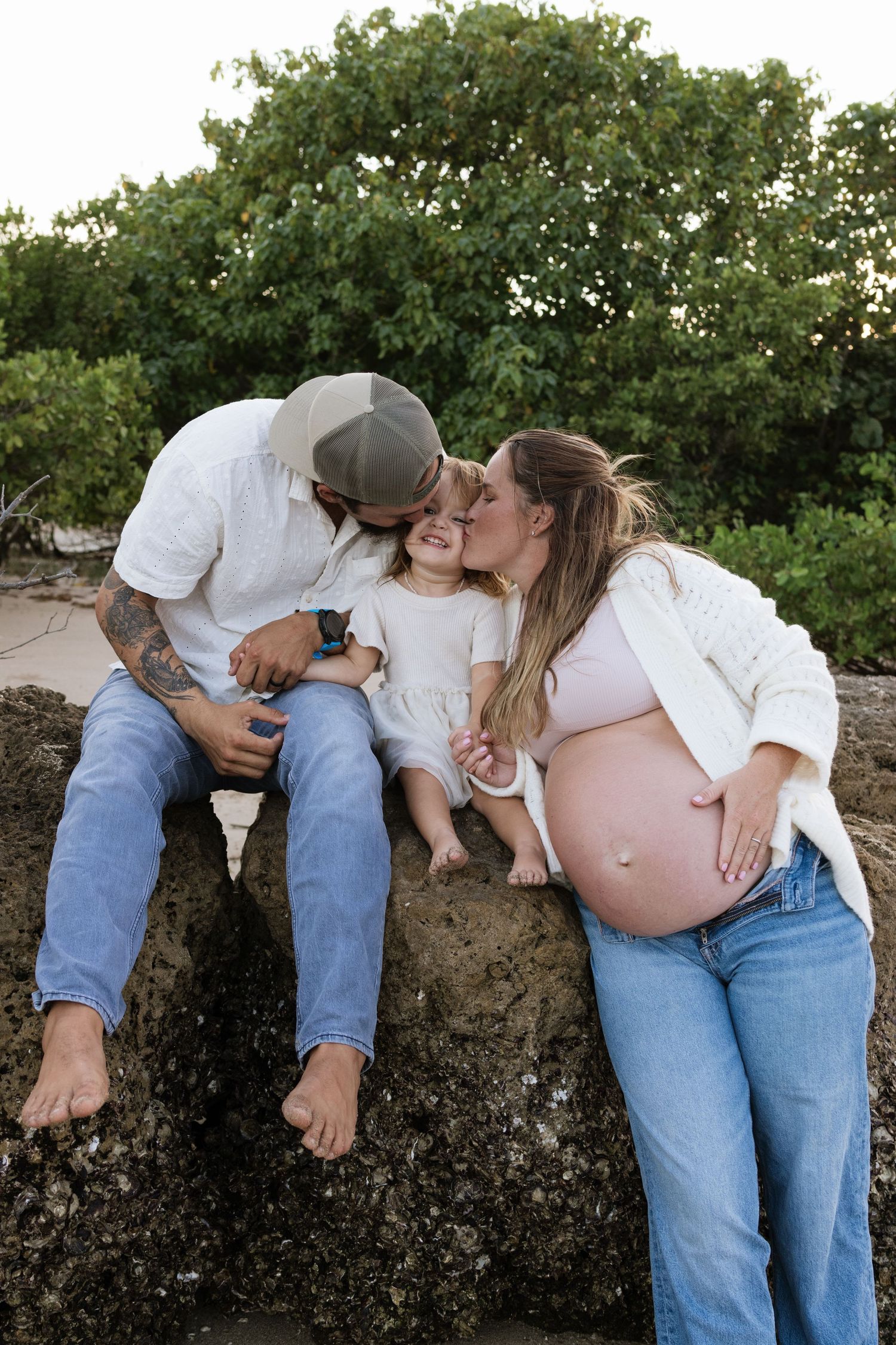 Intimate maternity photoshoot on a beach with greenery backdrop.