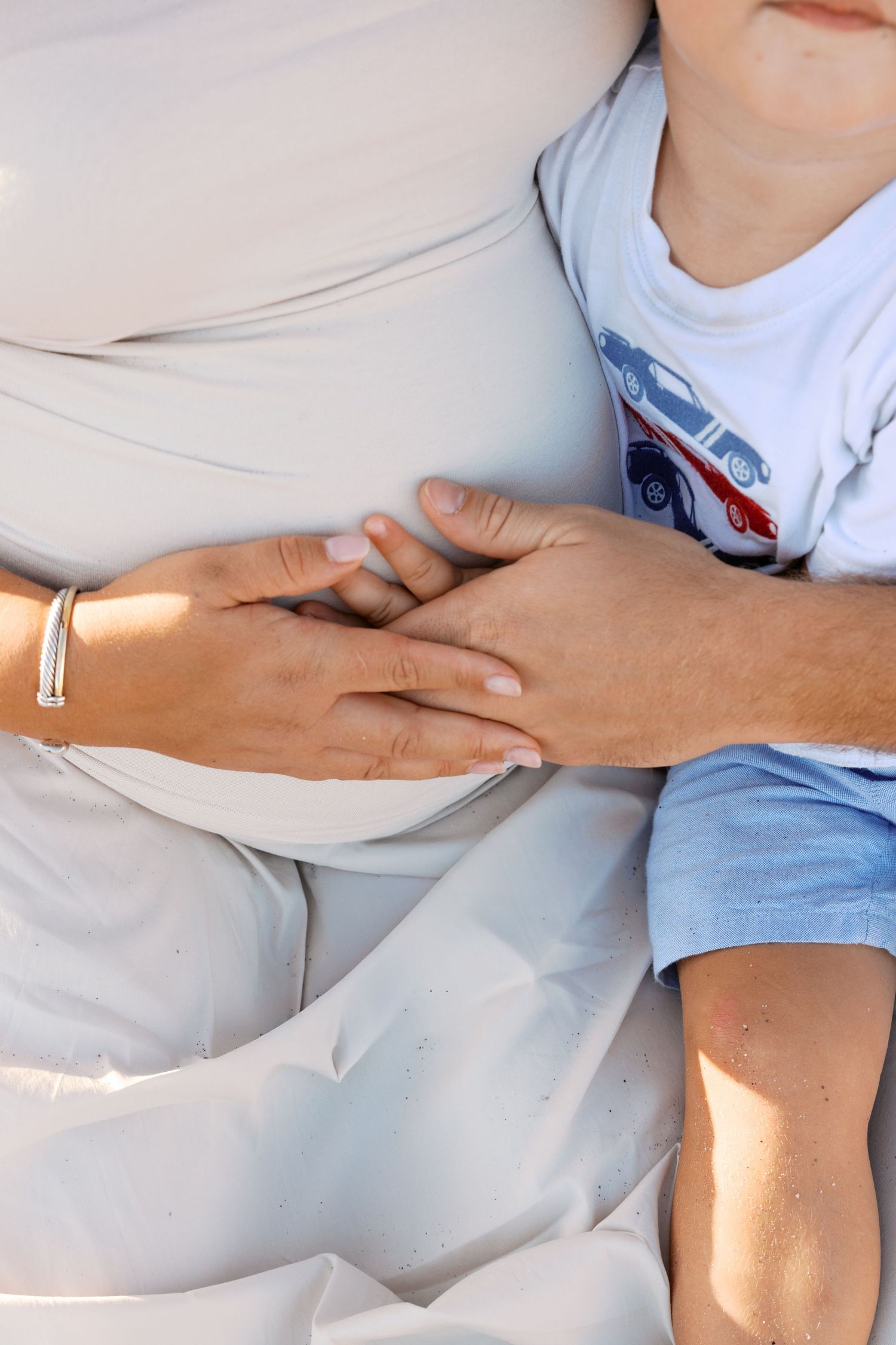A pregnant woman wearing a bracelet rests with a young child in a blue car shirt, their hands touching her baby bump.