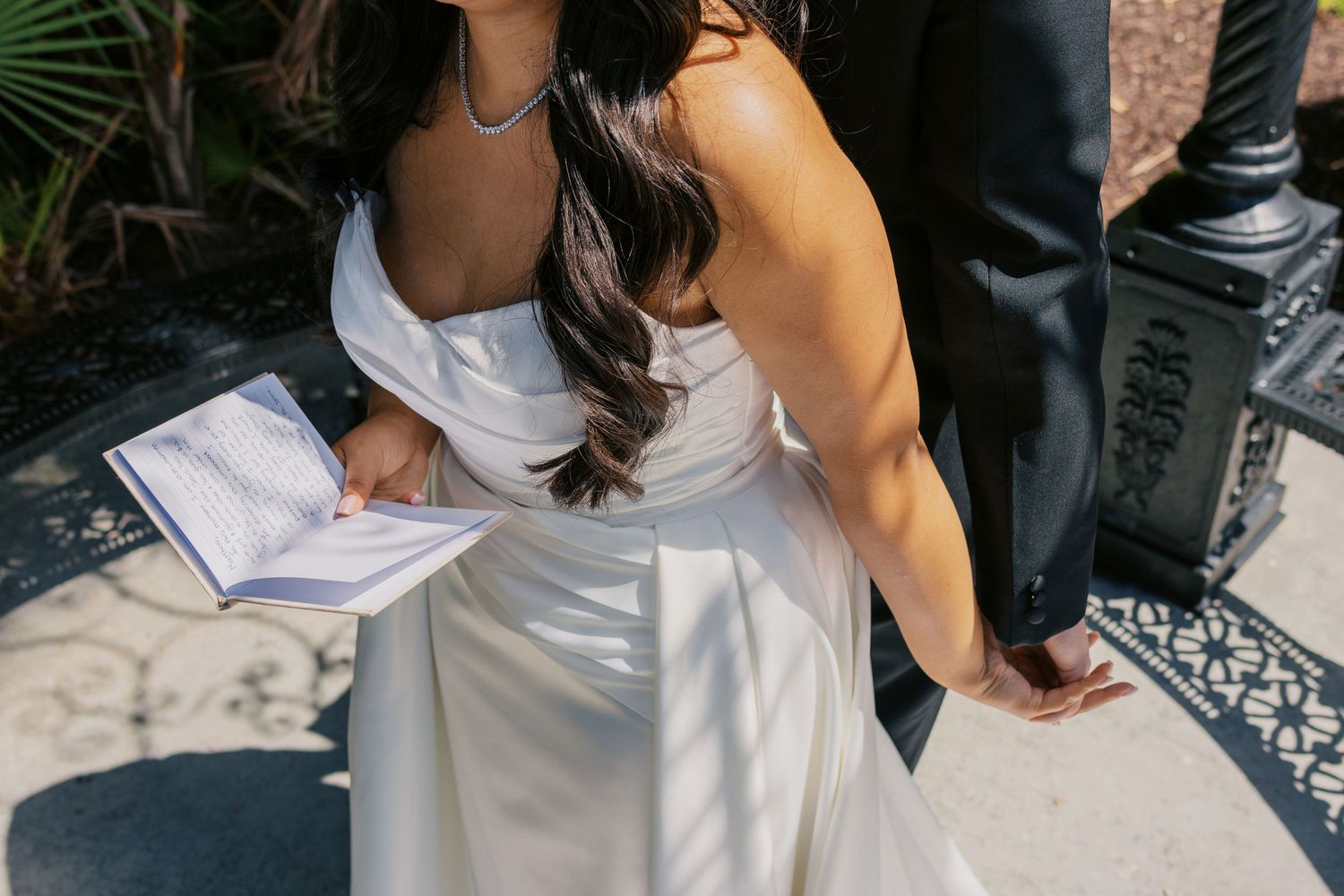 Bride in white strapless gown holding ceremony booklet, standing beside groom in black suit on decorative tile floor.