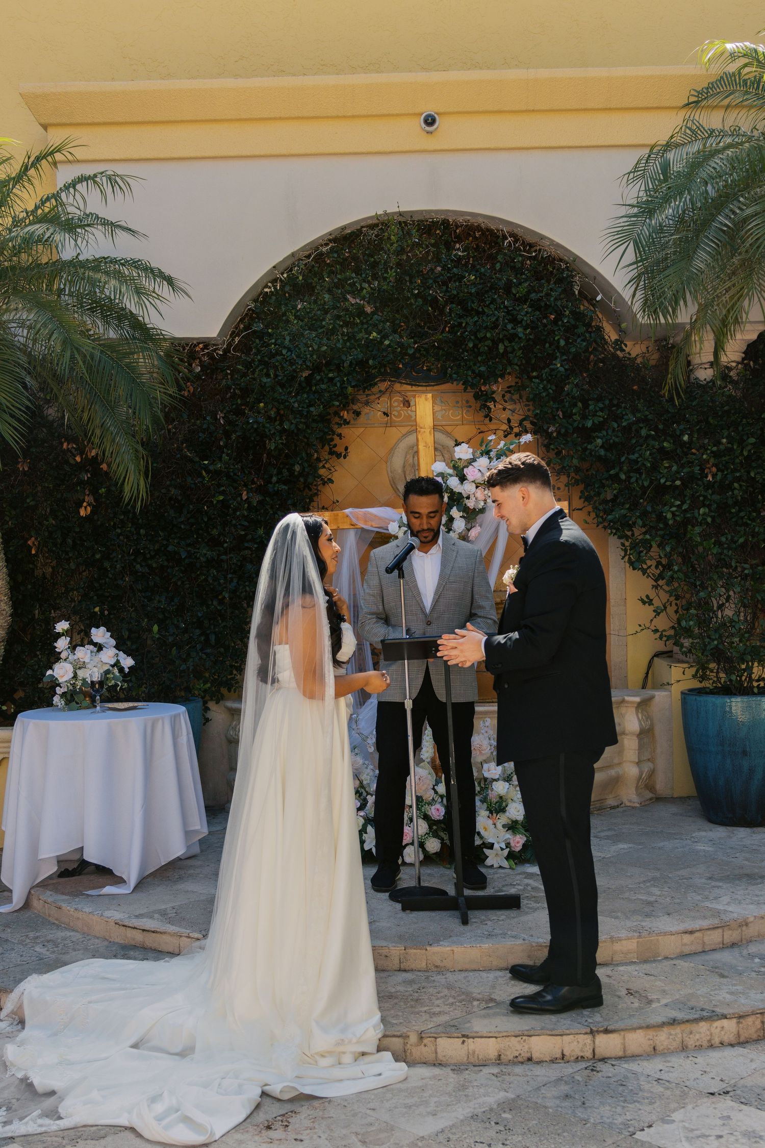 Bride and groom exchange vows outdoors at elegant wedding ceremony beneath ivy-covered arch with floral arrangements.