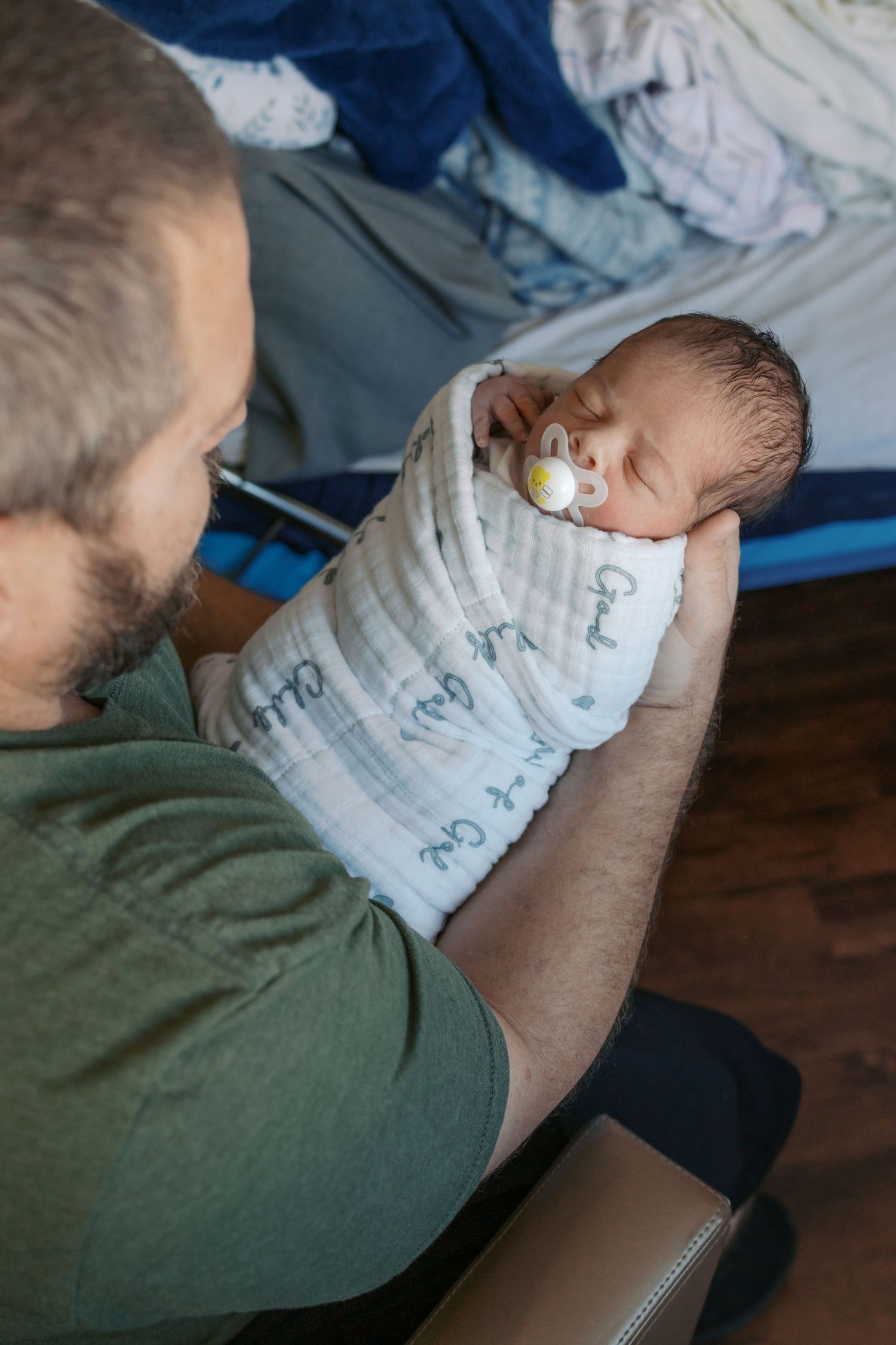 Father in green shirt holds swaddled newborn baby with pacifier, gazing lovingly at infant wrapped in white muslin blanket.