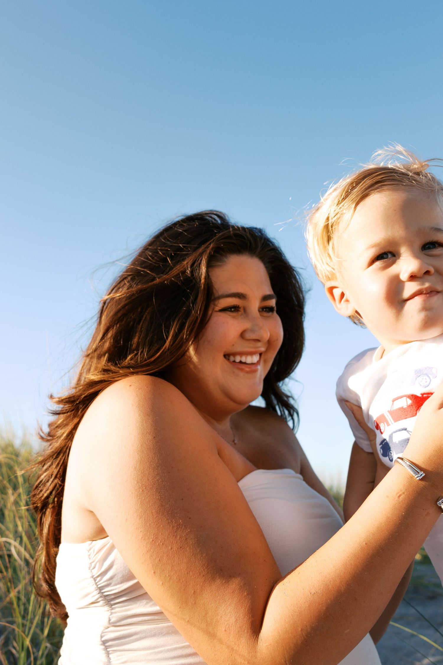 A smiling woman in a white top holds a young blonde toddler on a sunny beach with clear blue skies.