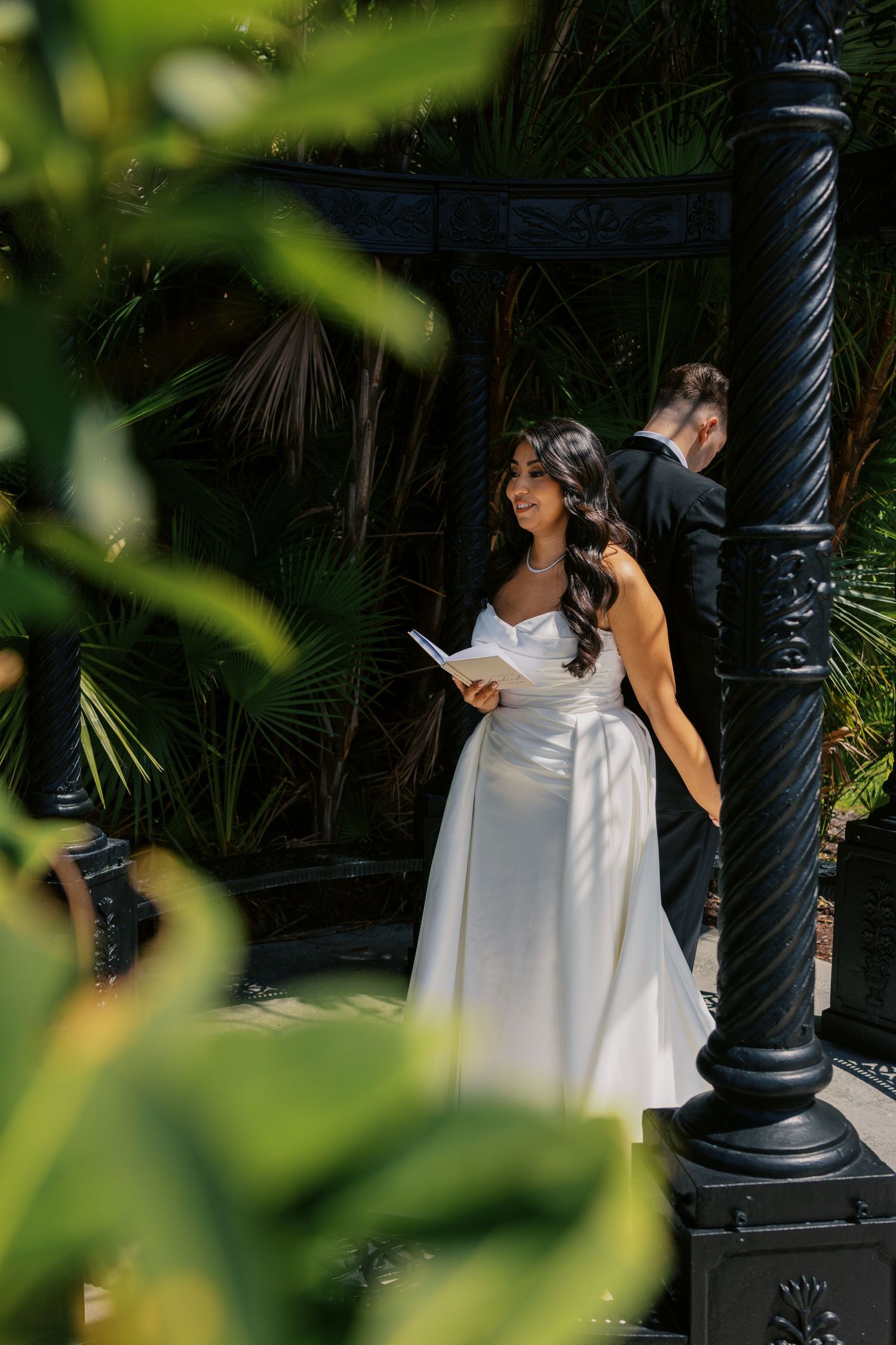 Bride in white satin gown holds vows booklet outdoors near black ornate gazebo pillars with lush tropical greenery.