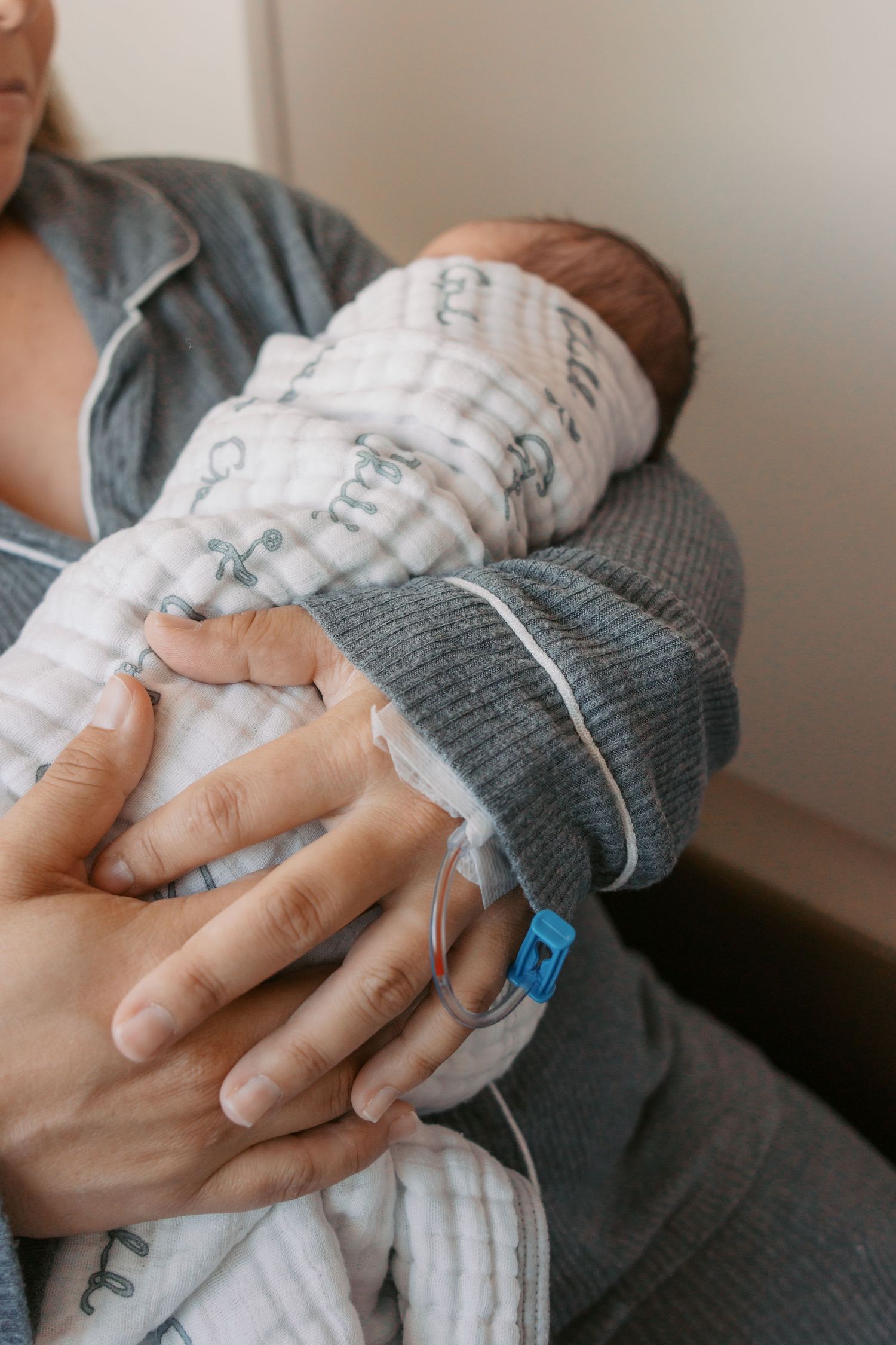 Newborn baby swaddled in white blanket being held by adult hands in a hospital setting.
