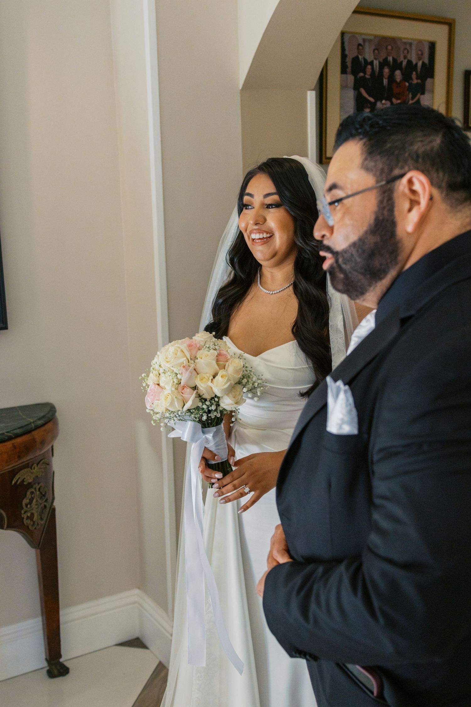 Smiling bride in white gown holding pink bouquet stands beside bearded groom in black suit inside elegant venue.