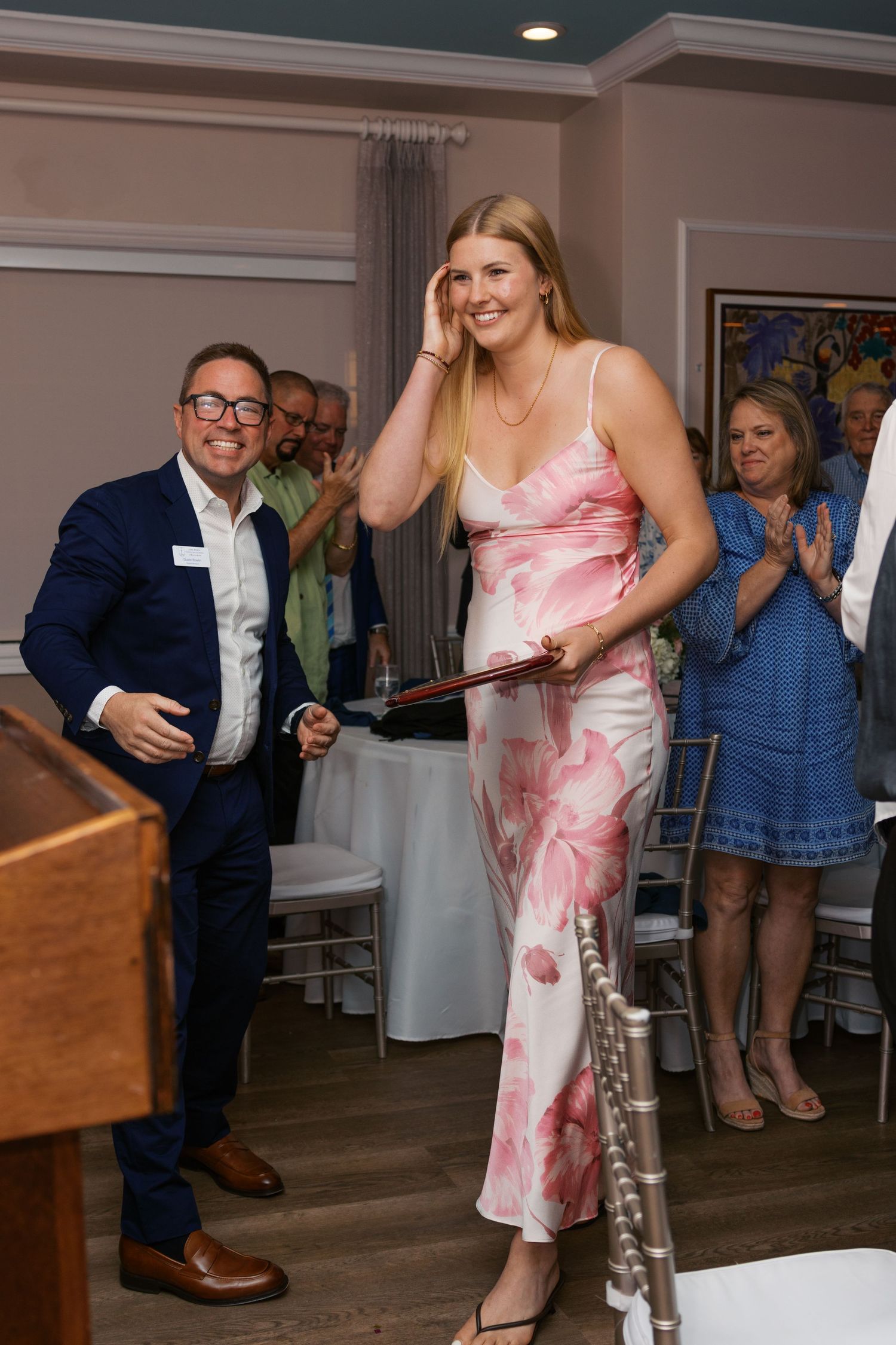 Woman in floral dress receives award at indoor ceremony while guests applaud in elegant dining room.