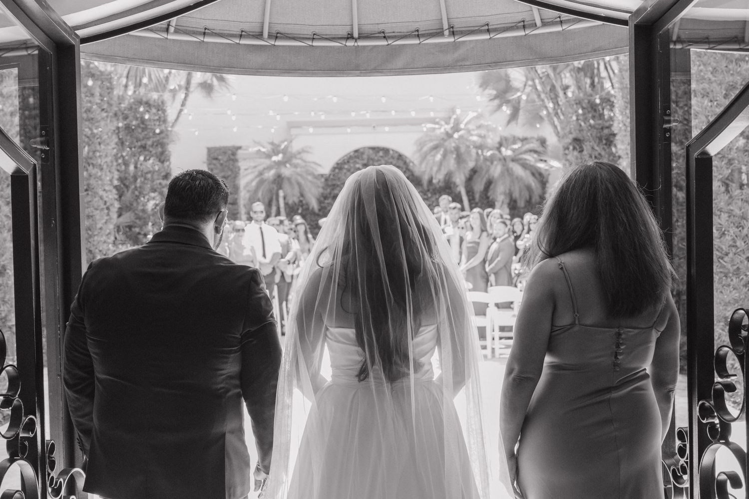 Black and white photo of a bride in veil flanked by two guests, about to walk down the aisle outdoors.