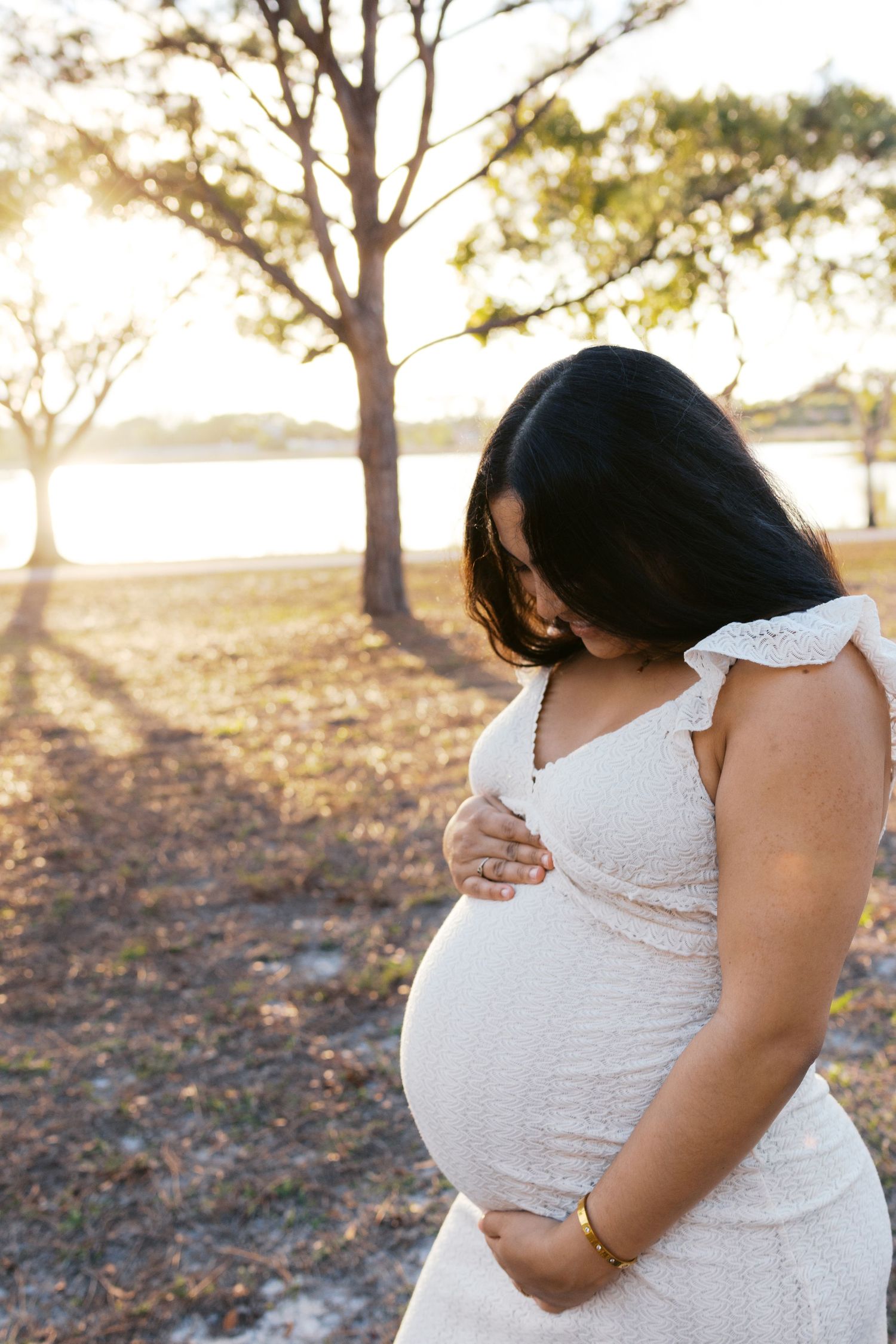 Pregnant woman in white lace dress holding baby bump in sunlit park at golden hour.