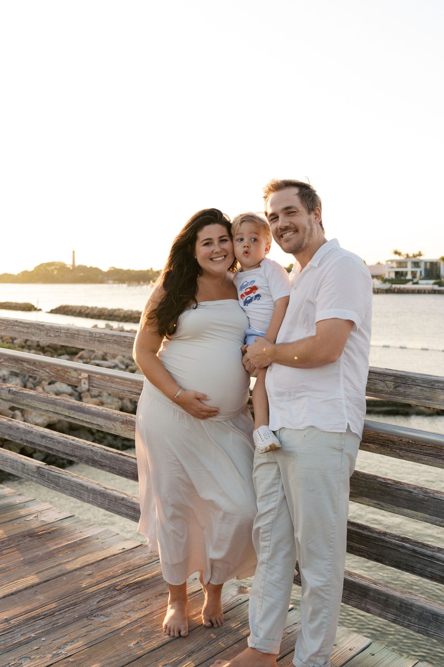 Smiling pregnant woman in white dress with husband holding toddler son on wooden boardwalk at golden sunset.
