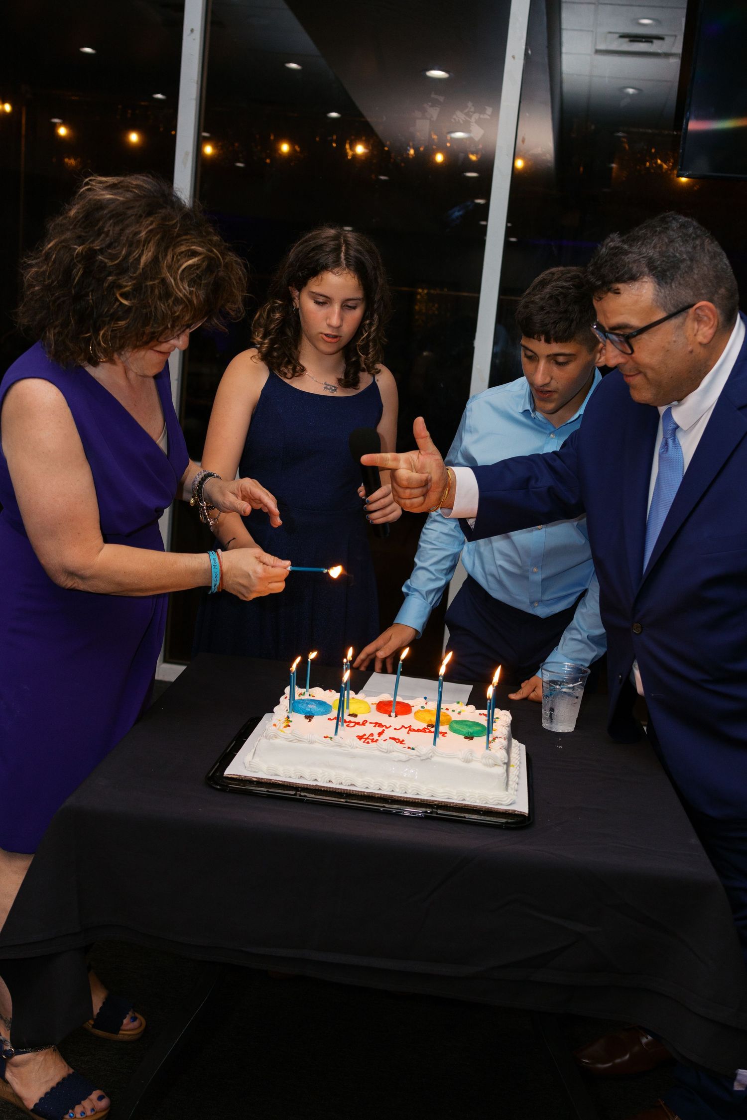 Family gathered around a white birthday cake with lit candles at a formal celebration event.