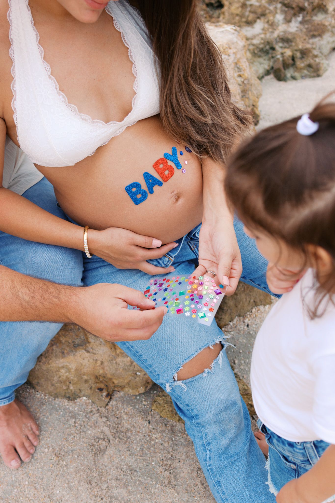 Pregnant woman with BABY letters on belly sits with child holding colorful stickers outdoors.
