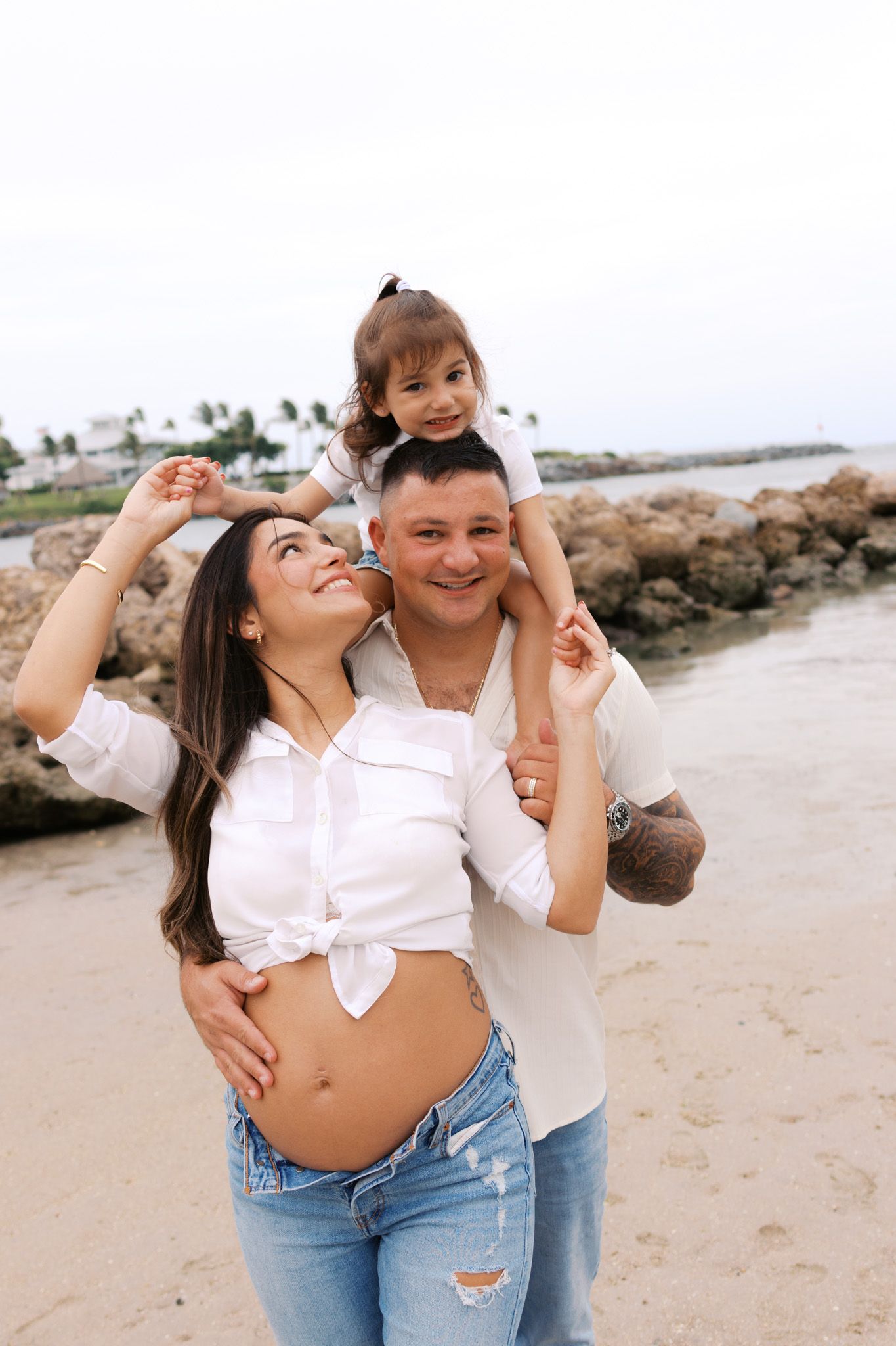 Happy pregnant family on beach, man carrying toddler on shoulders, woman in white crop top and jeans touching baby bump.