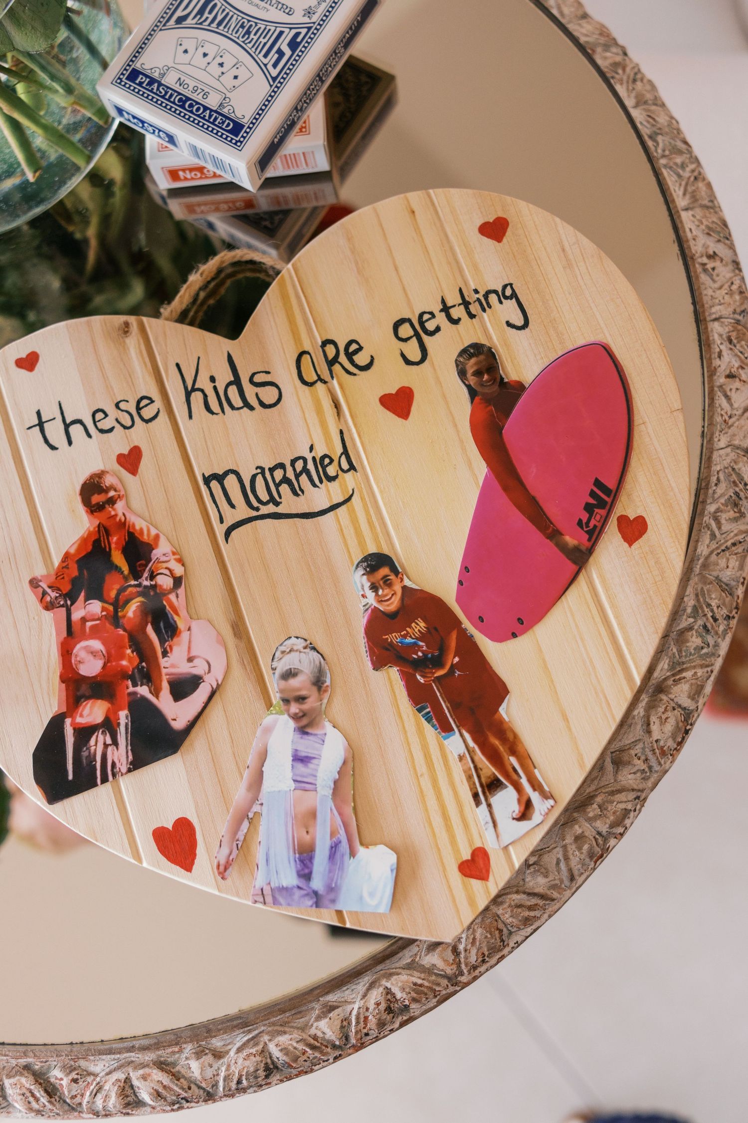 Heart-shaped wooden wedding sign with childhood photos reading these kids are getting married with red heart decorations.