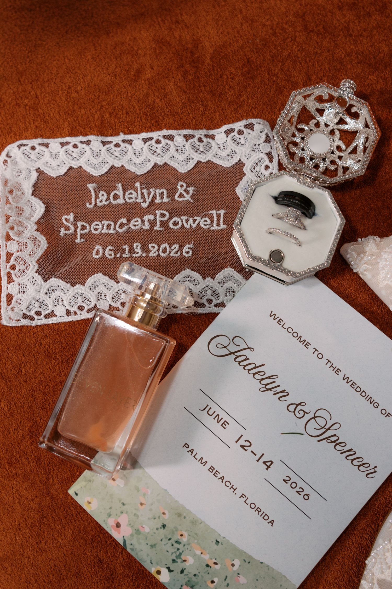 Wedding flat lay featuring Jadelyn and Spencer Powell June 13 2026 embroidered handkerchief, rings, perfume, and invitation.