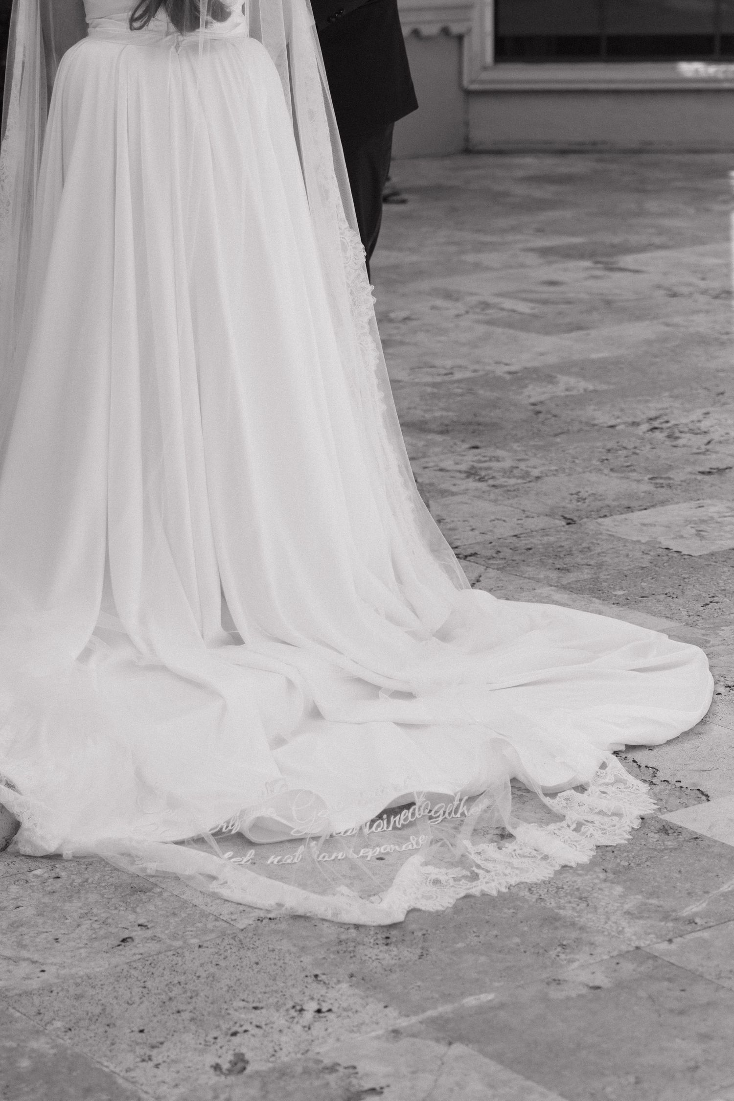 Black and white photo of a bride's flowing wedding dress train with lace trim on stone floor.