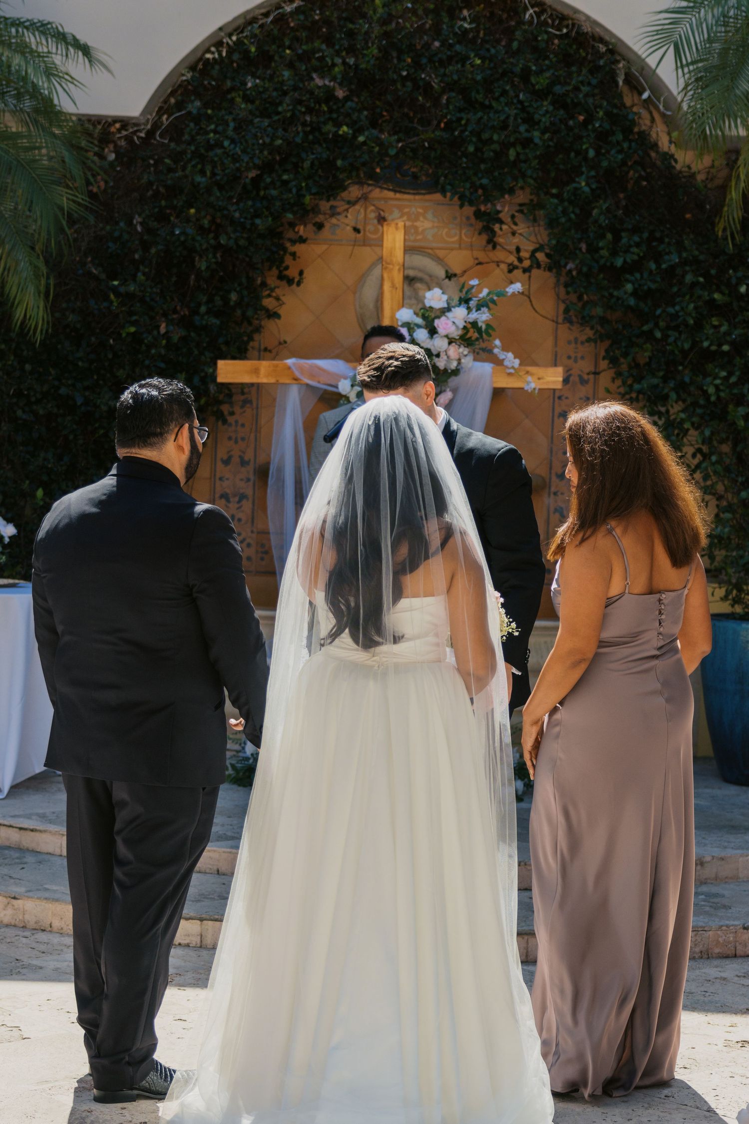 Bride in white gown and veil stands at outdoor wedding altar with cross, flanked by guests in formal attire.