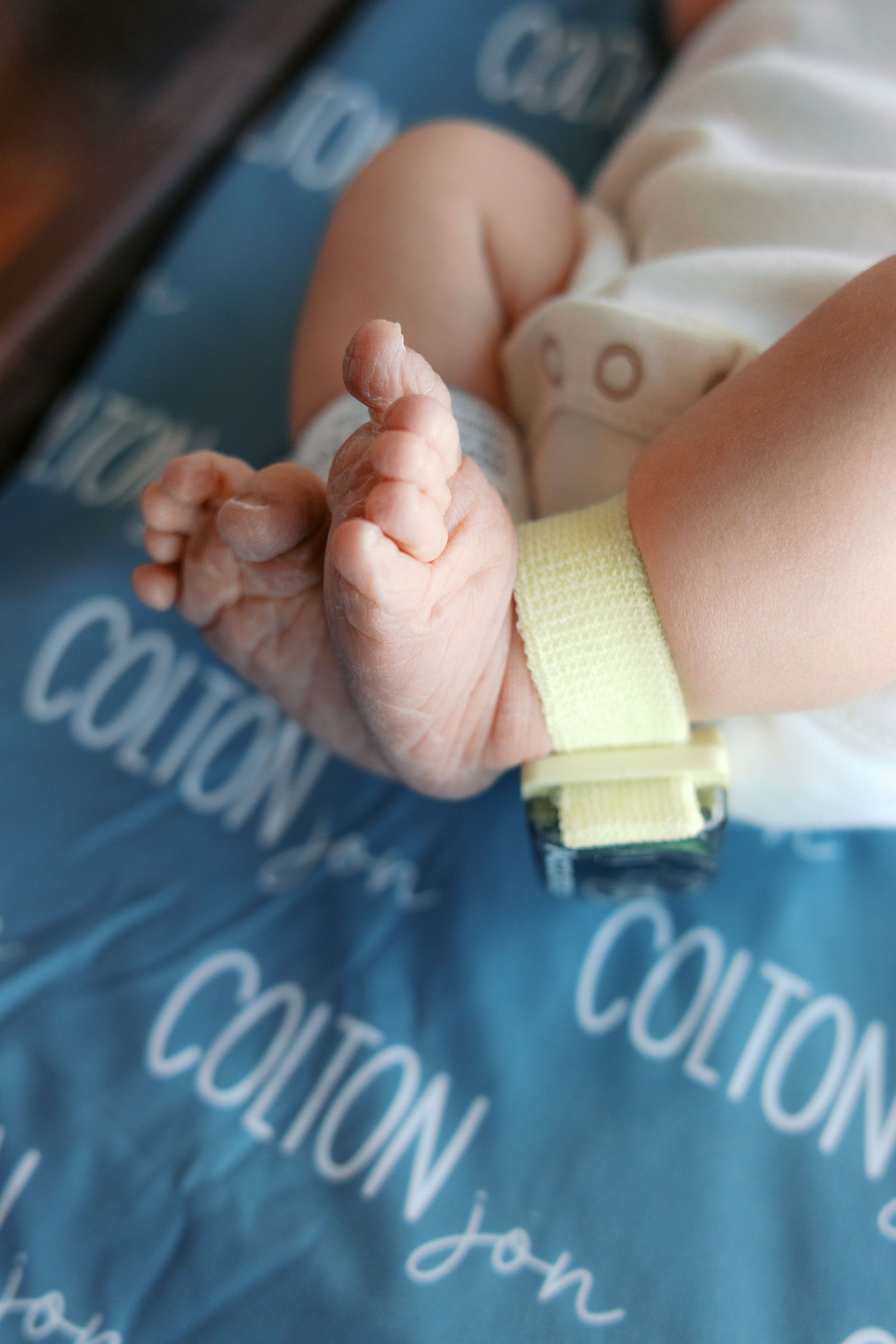 Newborn baby hands and feet with yellow hospital ID bracelet on blue blanket printed with name Colton Jon.
