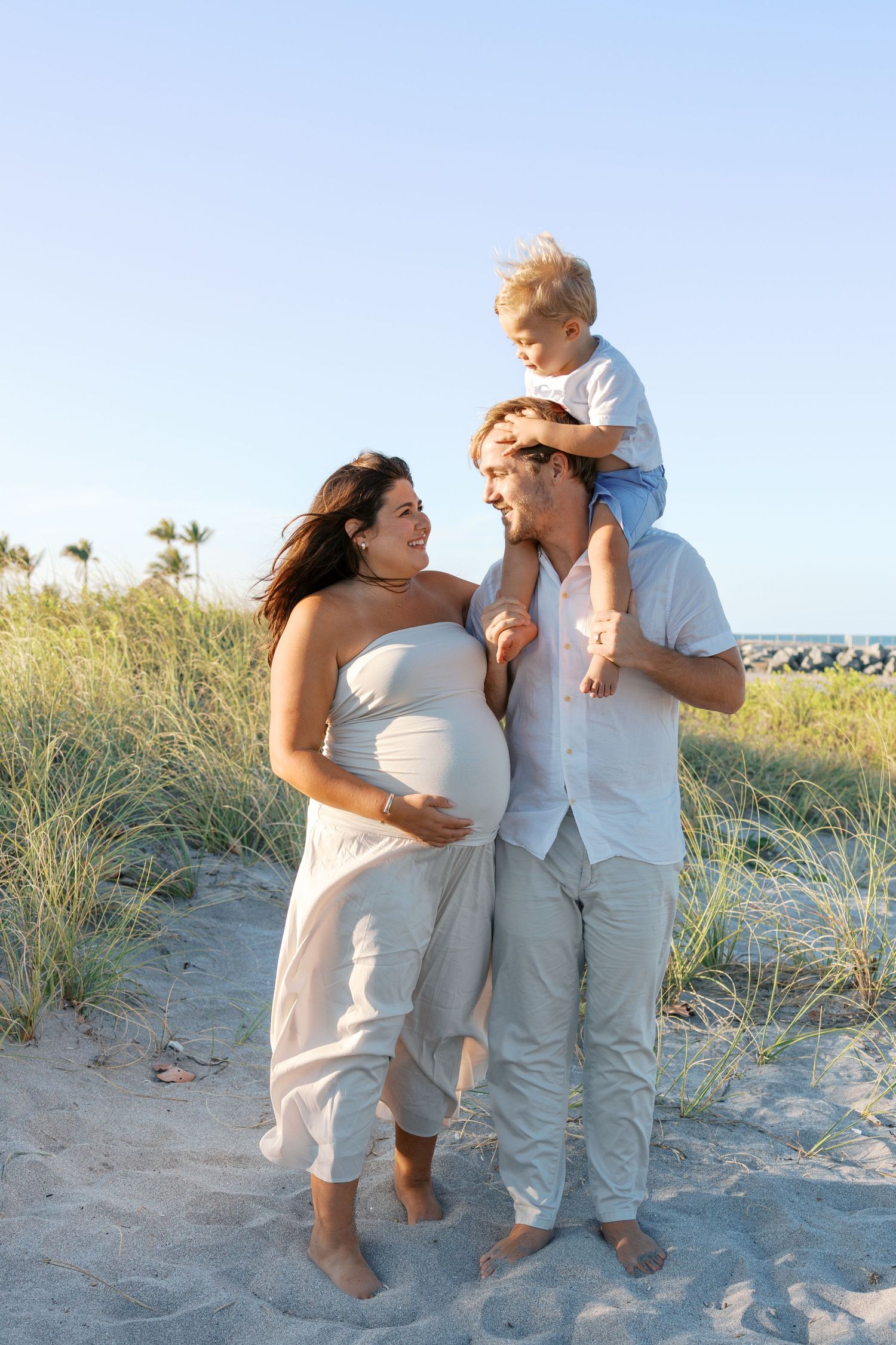 Pregnant woman and man with toddler on shoulders pose together on sandy beach in white outfits.