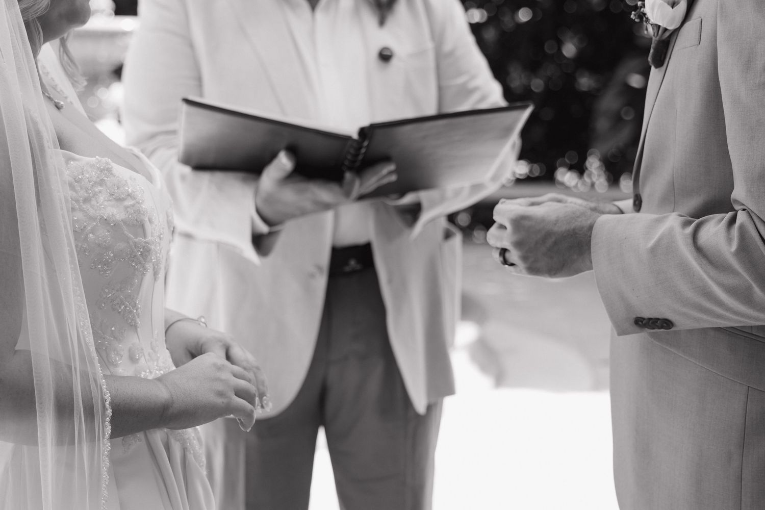 Black and white photo of a bride and groom exchanging vows with an officiant during an outdoor wedding ceremony.