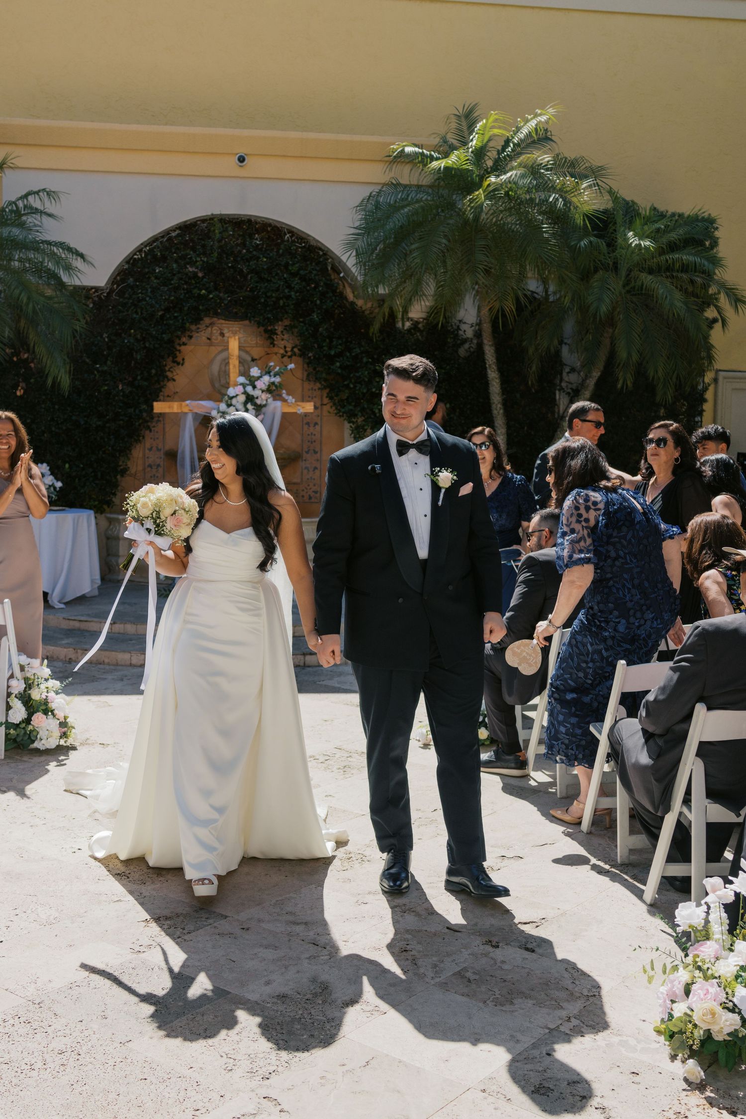 Bride and groom walk down the aisle at an outdoor wedding ceremony with palm trees and floral arch backdrop.