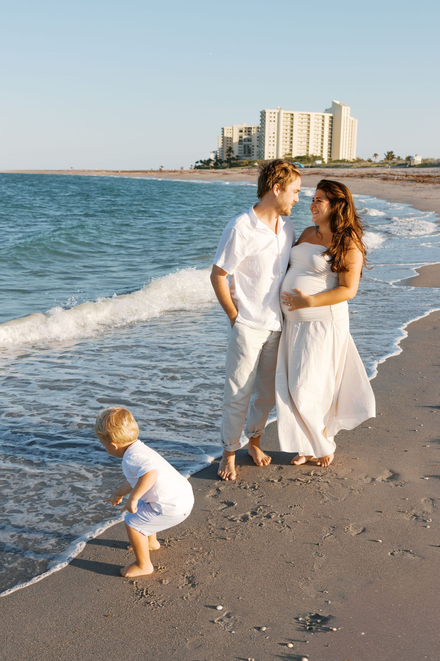 Pregnant woman in white dress with husband and toddler enjoying a sunny beach walk by the ocean.