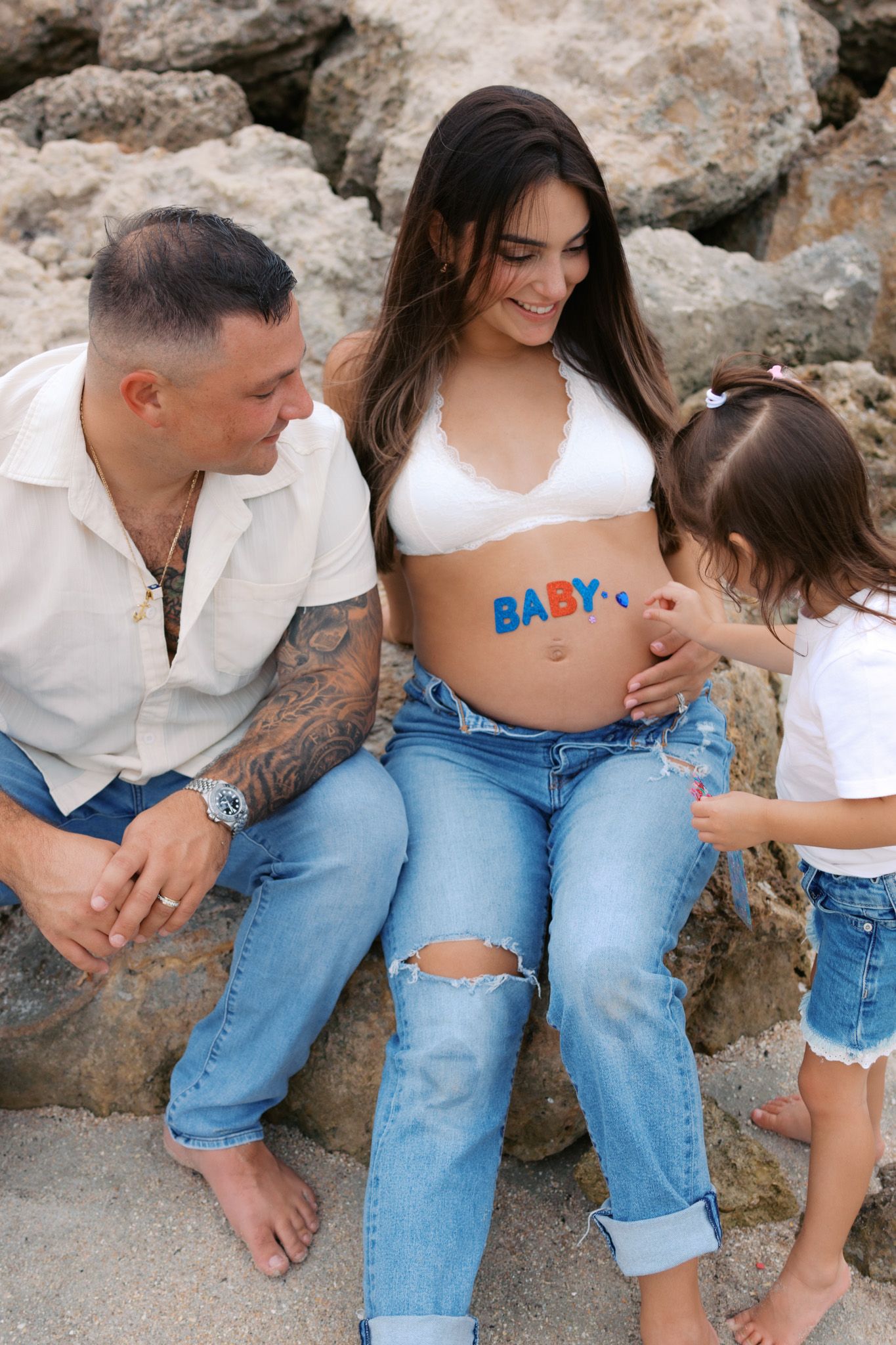 Pregnant woman with BABY painted on belly sits with tattooed partner and toddler daughter on rocky beach.