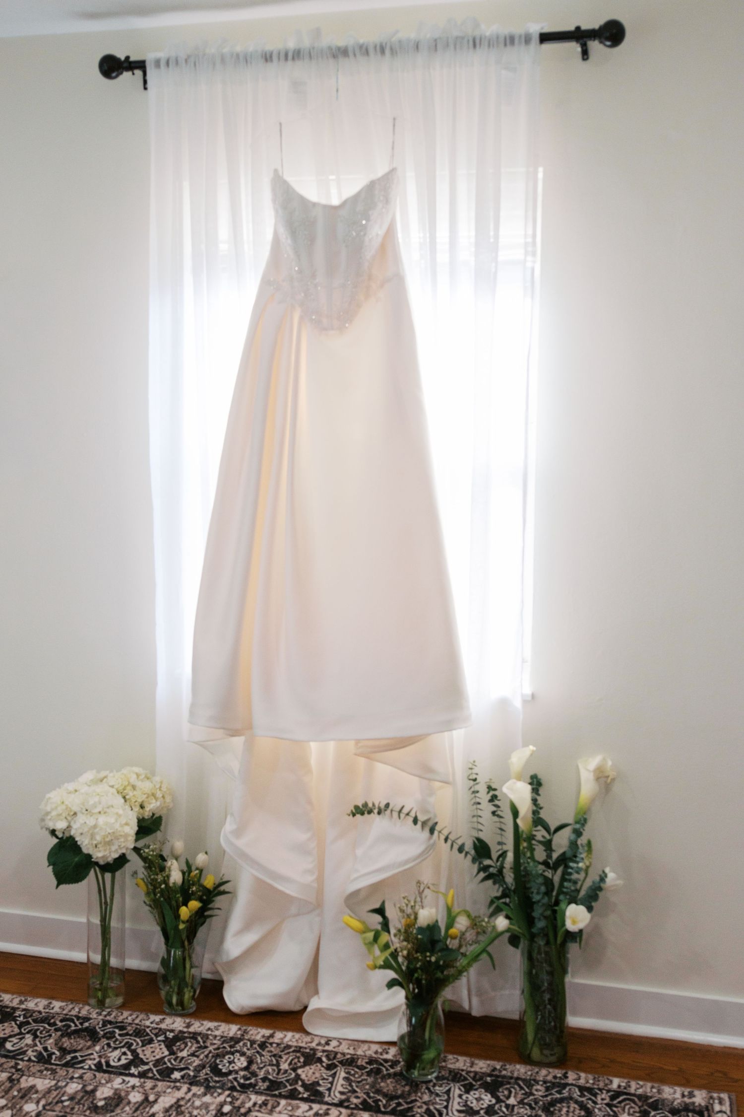A white strapless wedding gown hanging by a sunlit window, surrounded by elegant floral arrangements in glass vases.